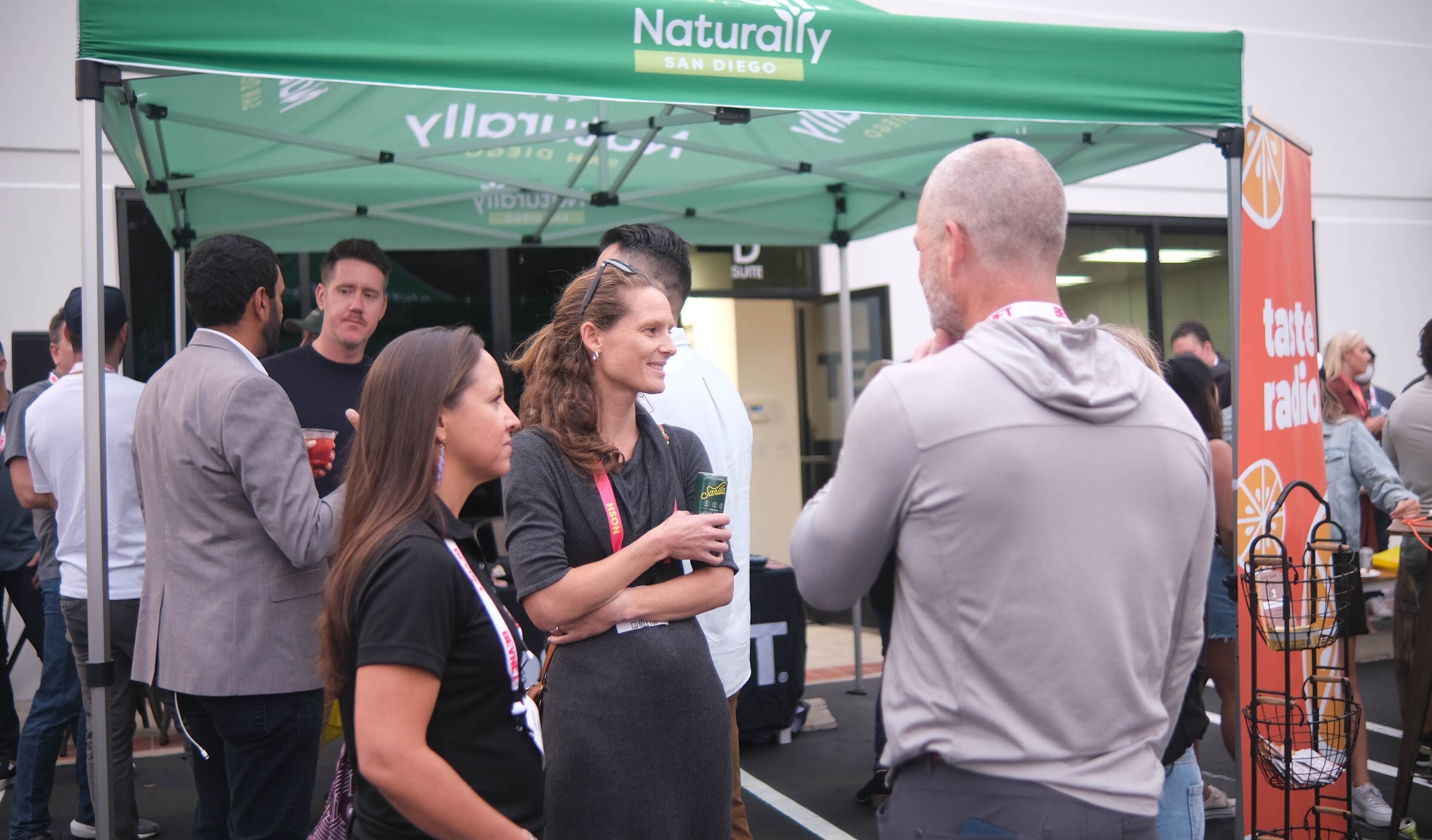 Guests in lanyards talk under a green Naturally San Diego tent with an orange Taste Radio banner nearby.