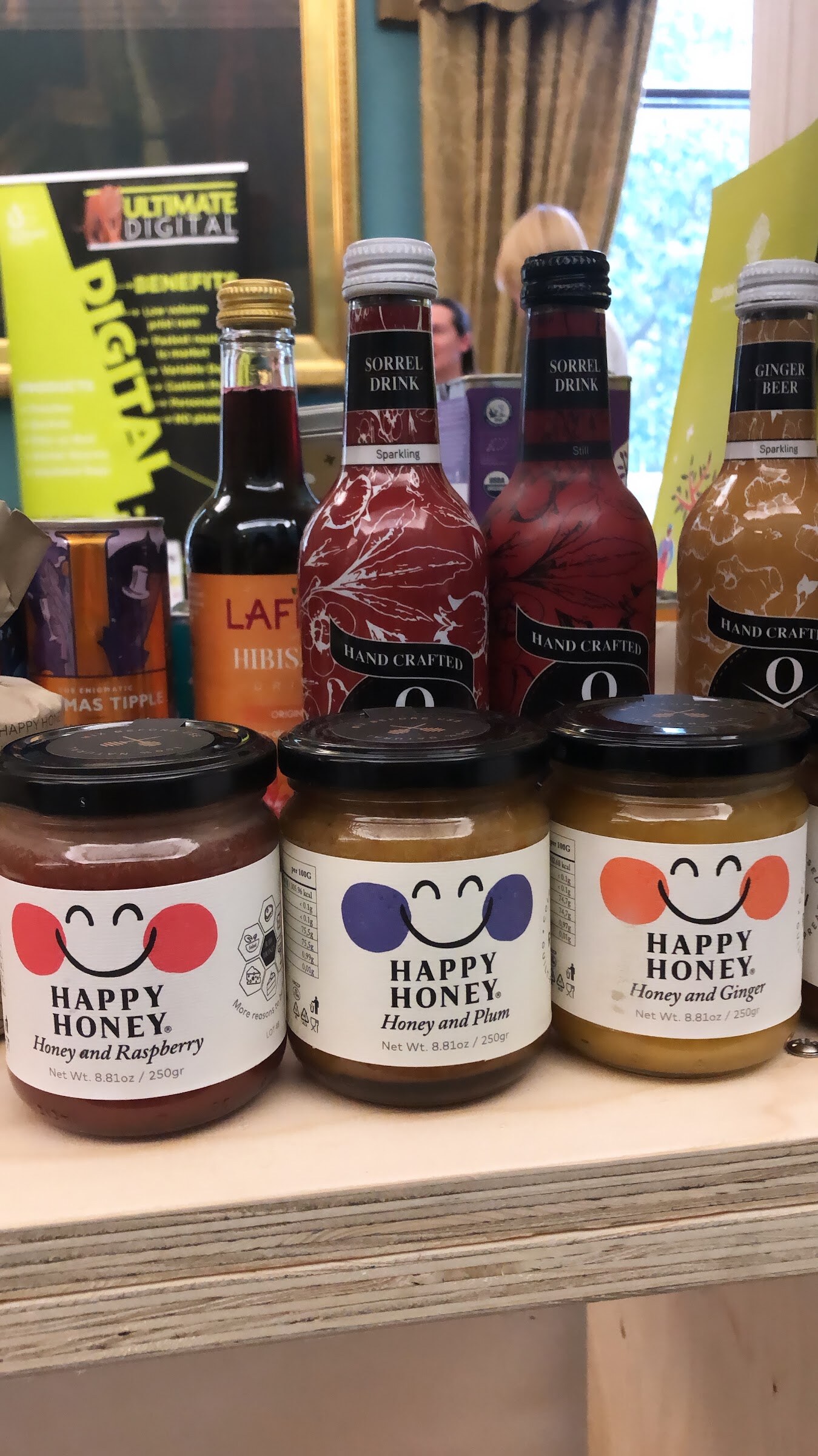 Three jars labeled Happy Honey and several bottles of a hand crafted drink on a wooden table.