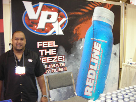 A man with a lanyard badge stands beside a VPX banner showing a large blue Redline bottle with the text 'Feel the freeze,' with cups stacked on a table nearby.