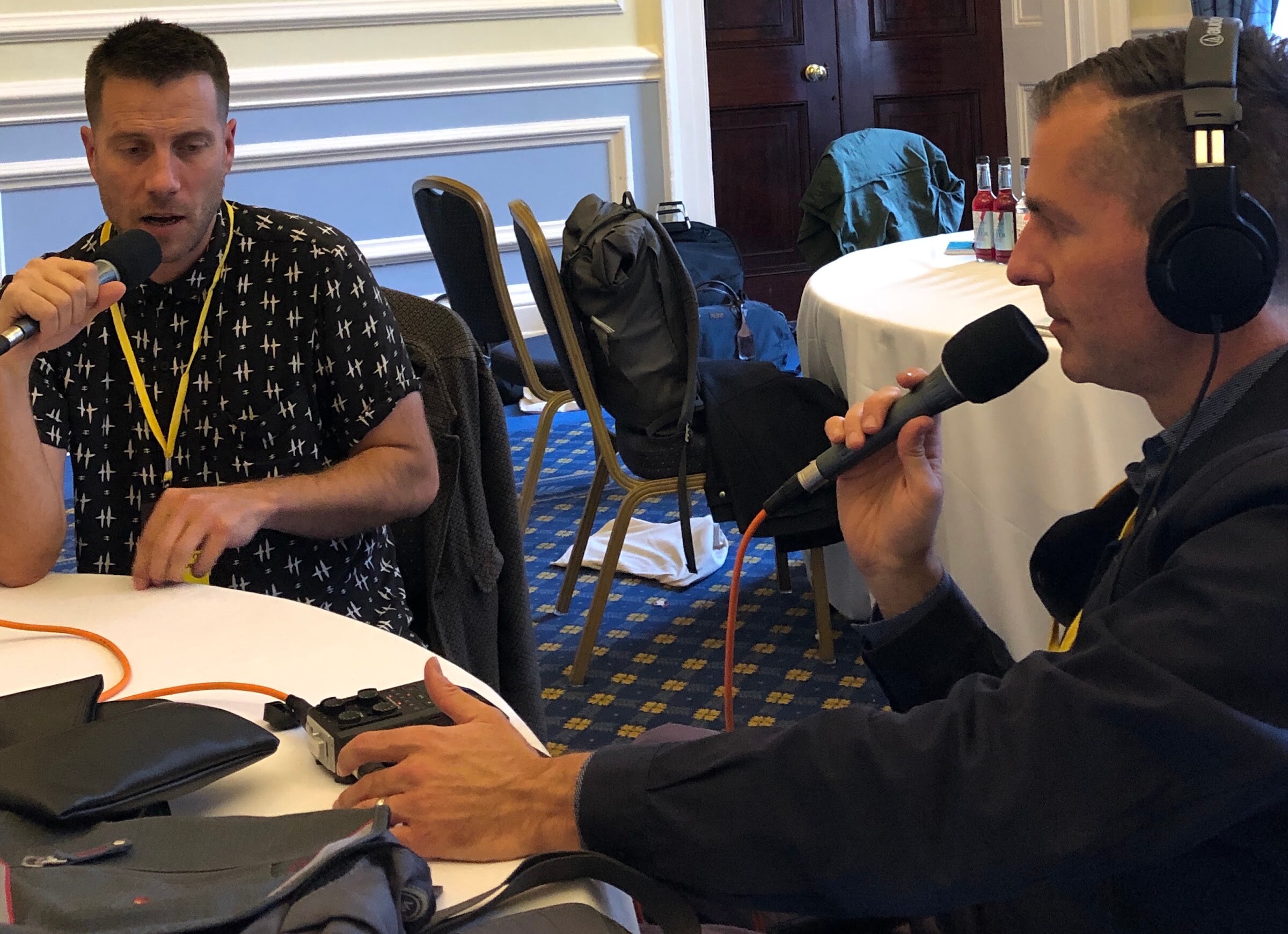 A man in a patterned shirt with a lanyard and a man in headphones speaking into microphones at a table with a recorder.