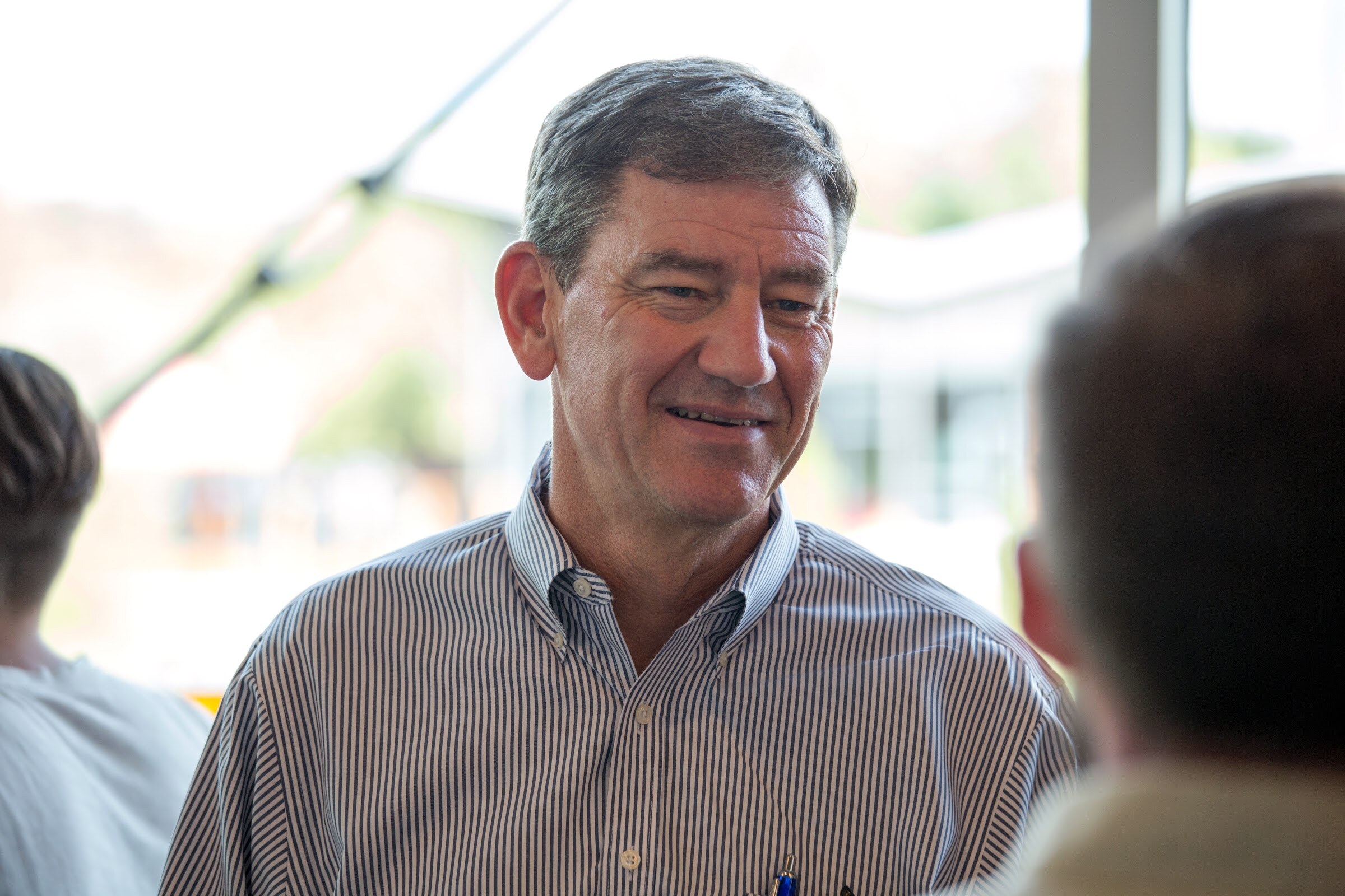A gray-haired man in a striped shirt faces another attendee near a bright window.