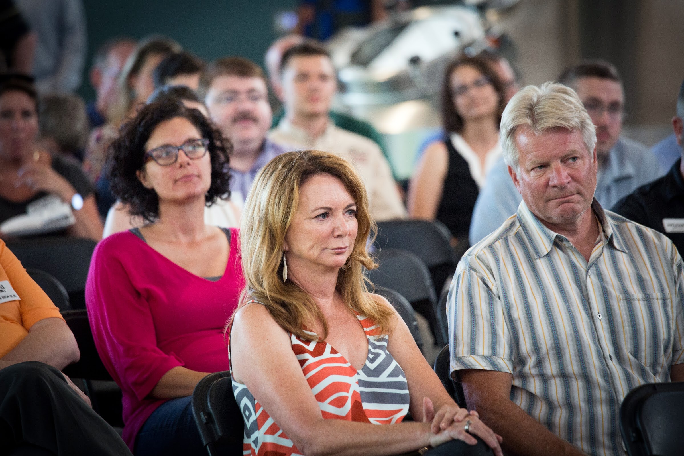 A woman in a patterned top and a man in a striped shirt sit in front of other audience members.