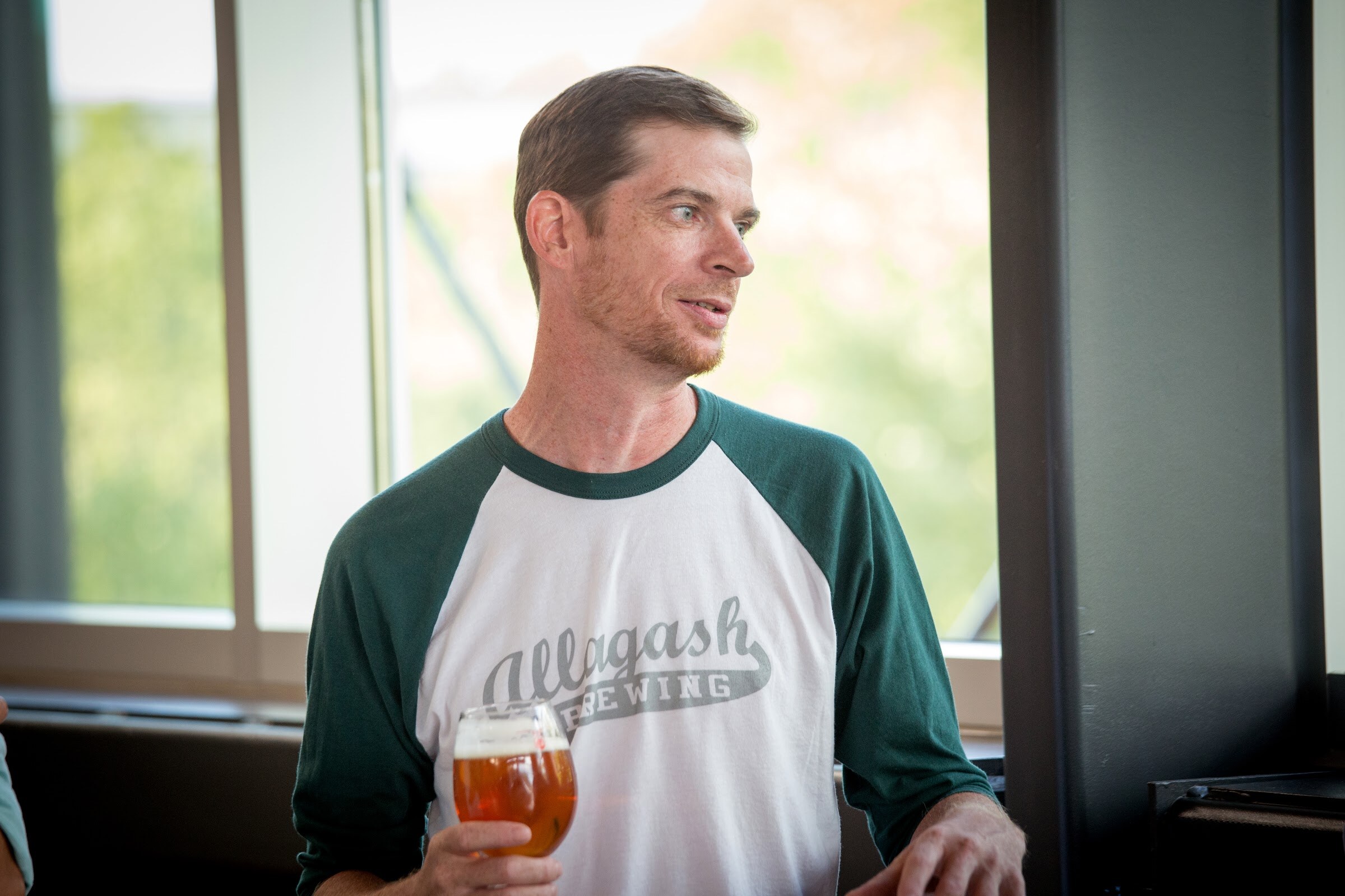 A man in an Allagash Brewing shirt holds a stemmed beer glass while looking to his right.