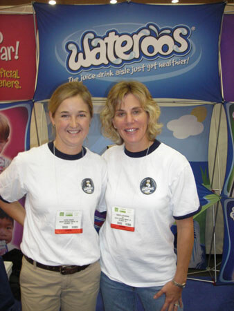 Two smiling women in white t-shirts and badges stand side by side in front of a blue Wateroos banner reading 'The juice drink aisle just got healthier!'