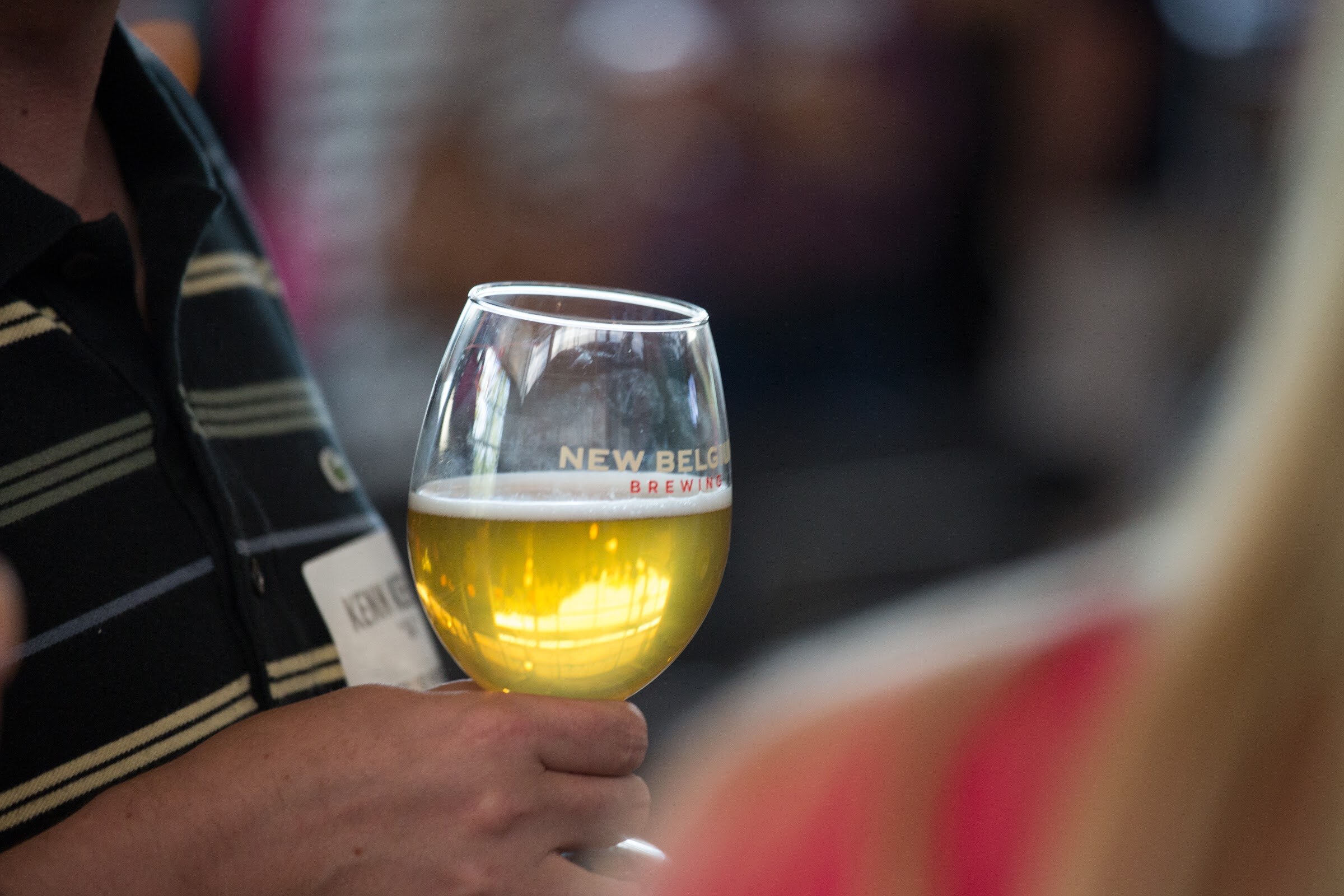 A hand holds a stemmed glass of golden beer with New Belgium Brewing lettering.