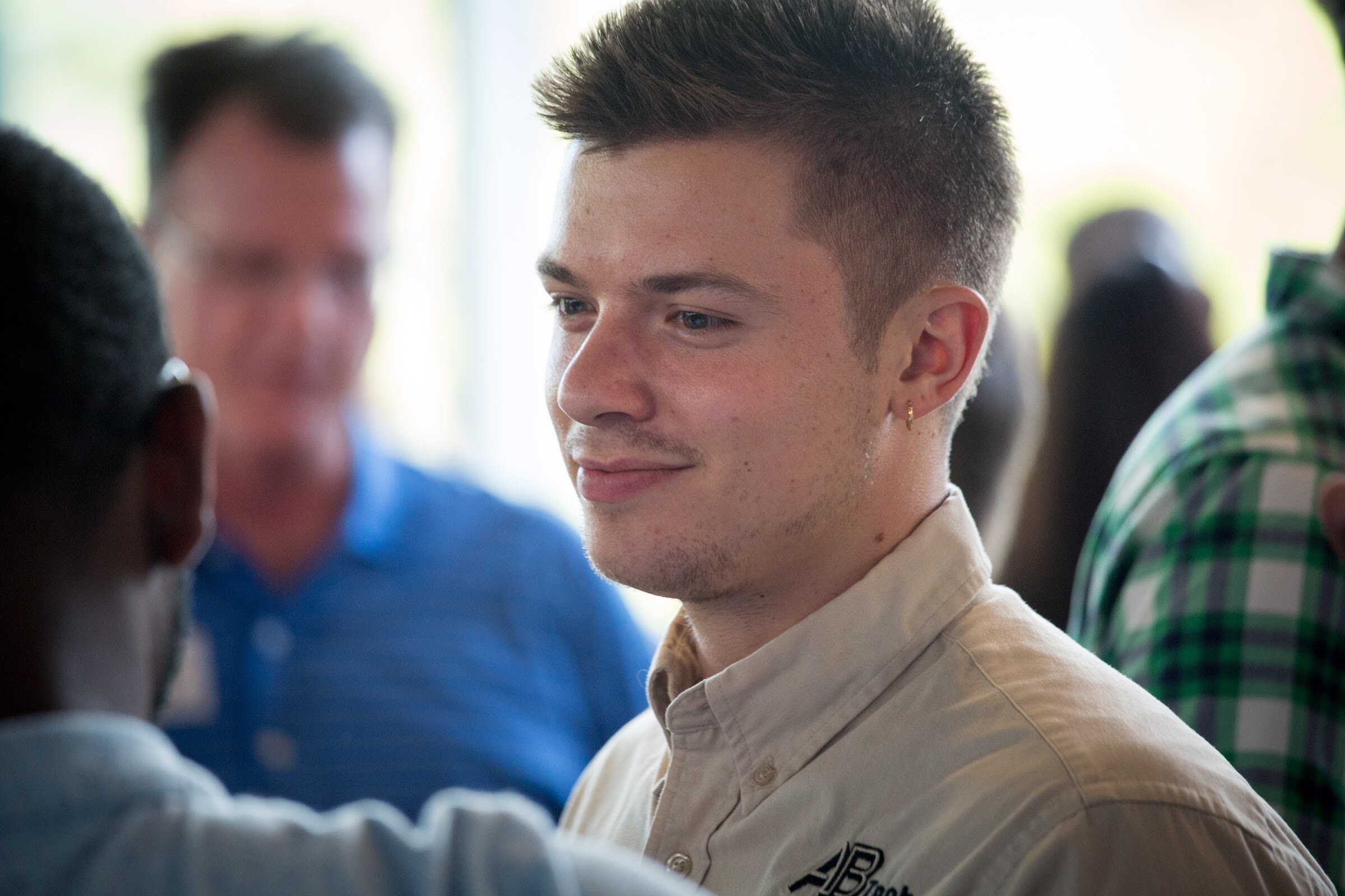 A young man in a beige button-down shirt faces another attendee, with guests behind him.