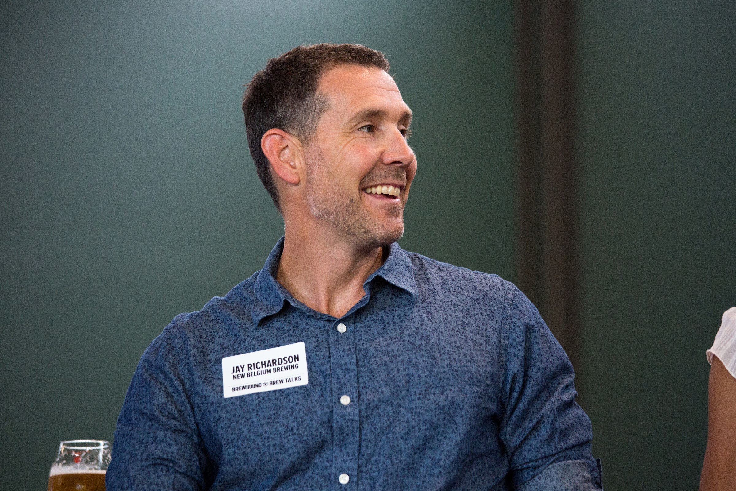 A man in a blue patterned shirt smiles to his right against a green wall.