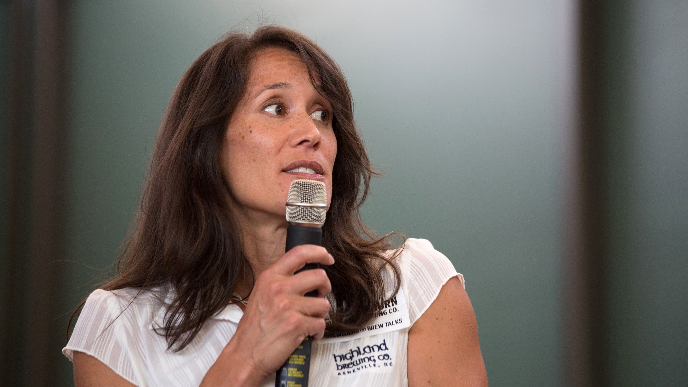 A woman in a white blouse holds a microphone and looks to her left.