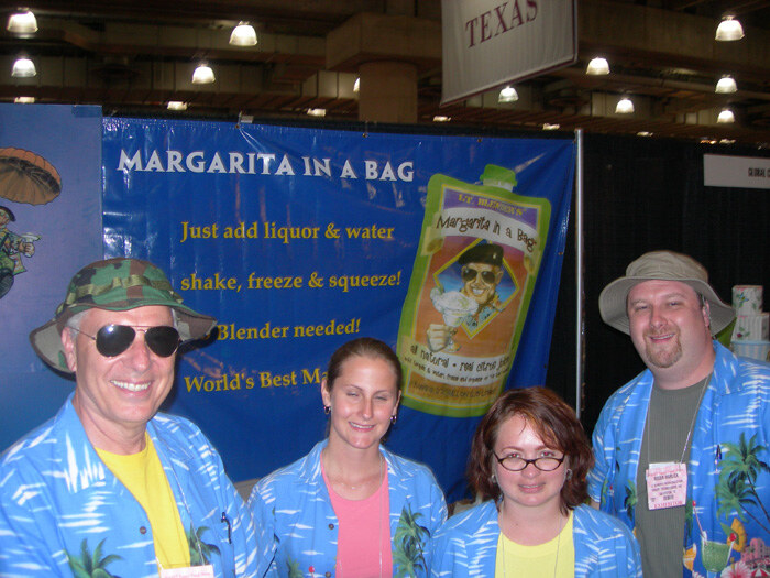 Four people in blue Hawaiian-style shirts stand in front of a blue Lt. Blender's Margarita in a Bag banner that reads 'Just add liquor & water'.