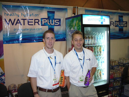 Two men in white polo shirts and badges hold Fuze bottles in front of a Water Plus banner and a lit refrigerated display case of drinks.