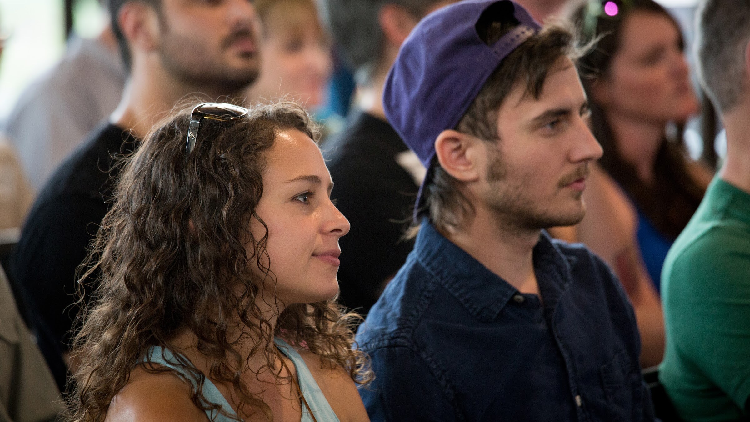 A woman with curly hair and a man in a backward purple cap look toward the discussion.