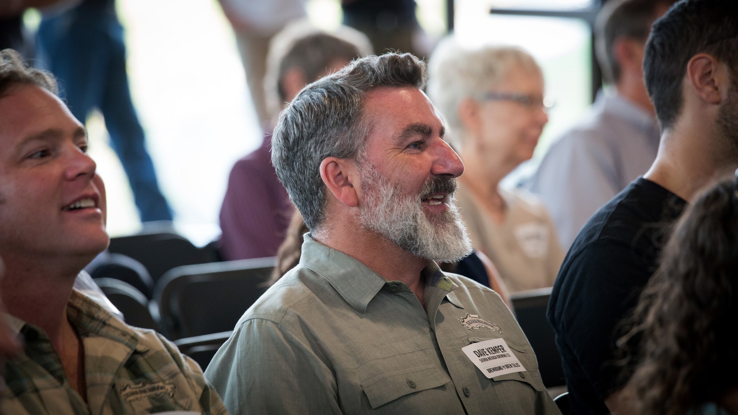 A gray-bearded man in a green shirt laughs while seated among other guests.