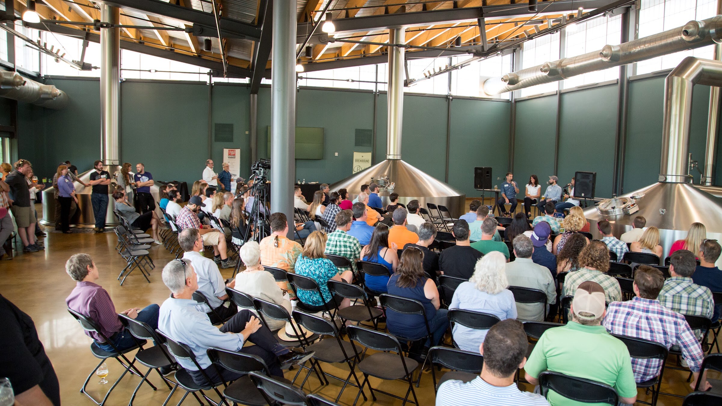 A wide view shows rows of attendees, large stainless-steel brewing tanks and four panel participants.