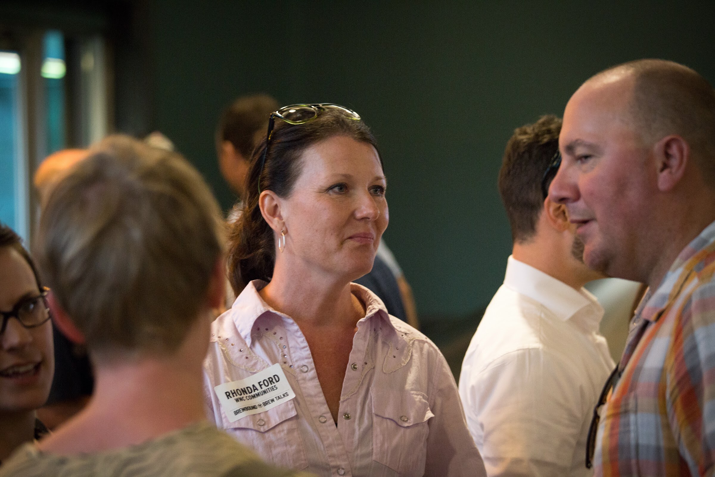 A woman in a pale pink shirt listens to a man in a plaid shirt amid other attendees.