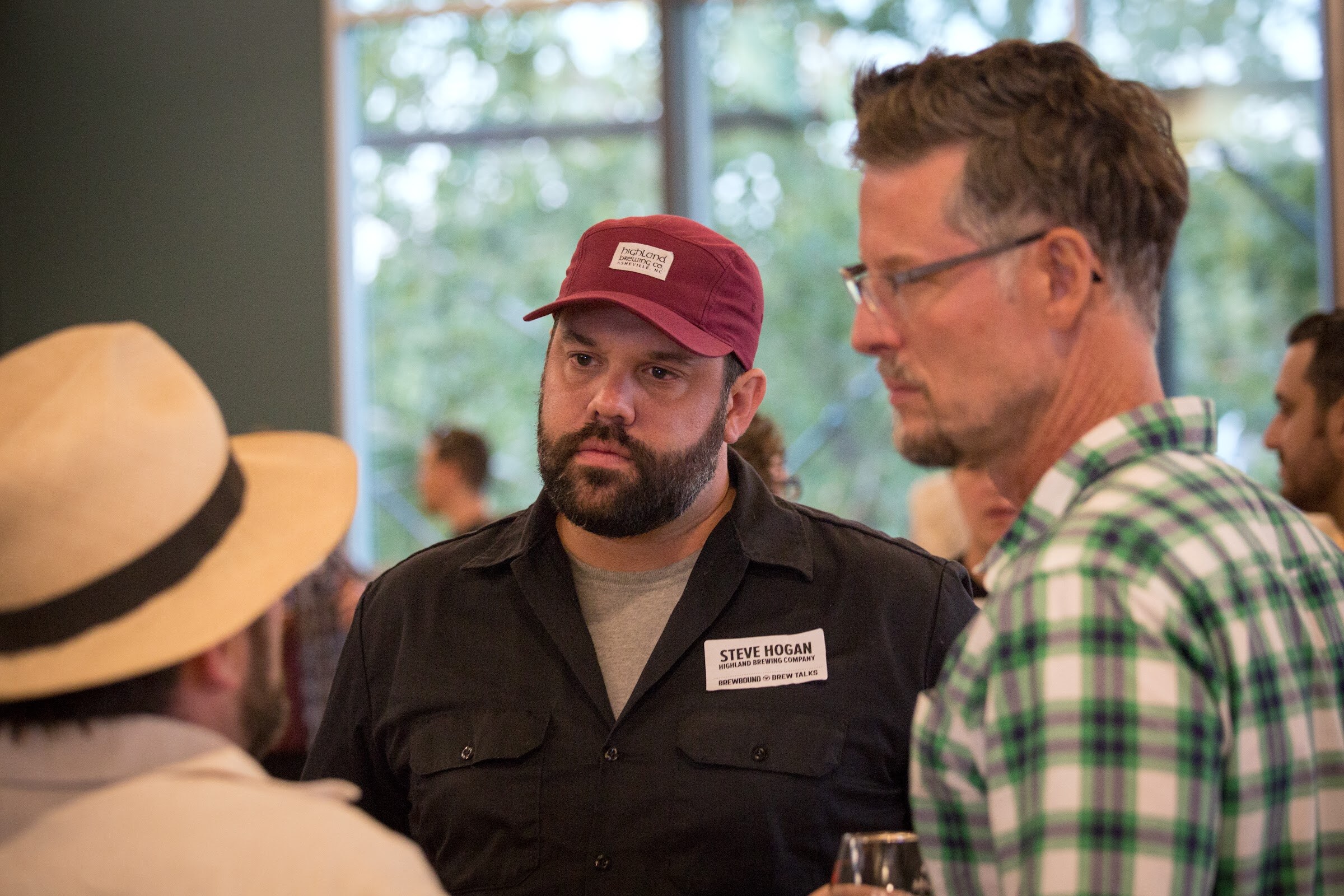 A bearded man in a red cap and a man in a green checked shirt face another guest wearing a light hat.