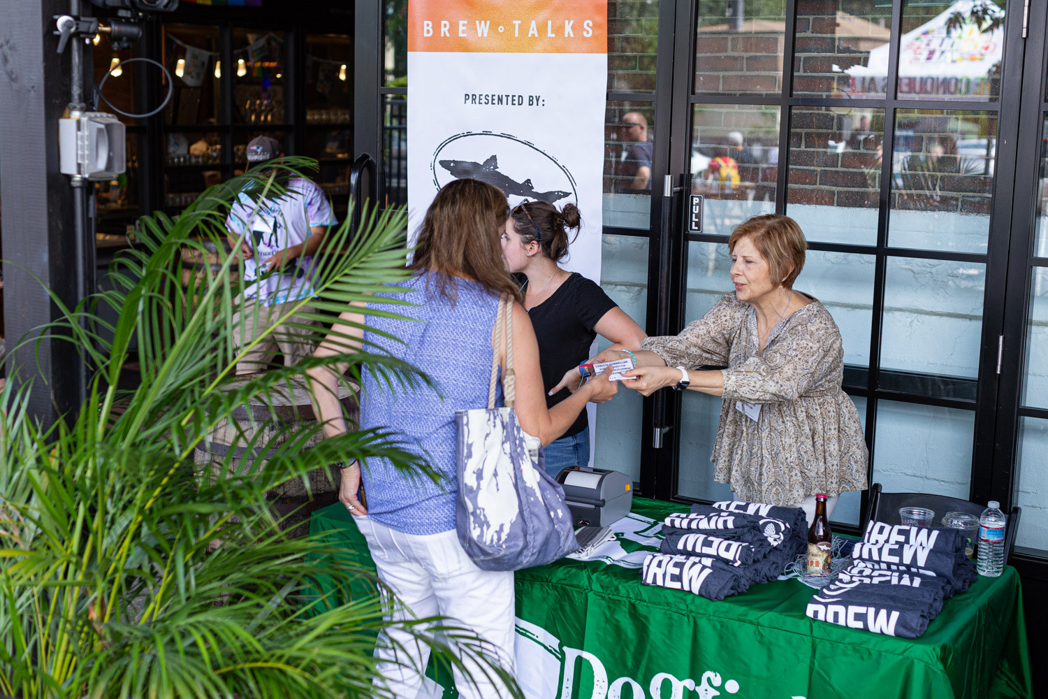 A woman in a blue top and white pants takes a badge from a woman in a paisley blouse behind a green table stacked with navy Brew shirts, beside a Brew Talks banner presented by Dogfish Head and palm fronds.
