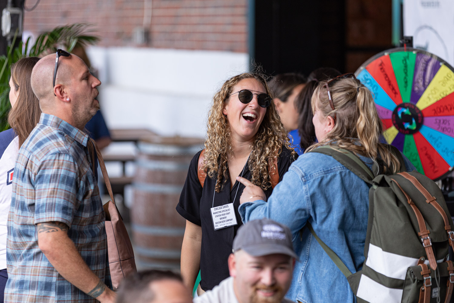 A laughing woman with curly hair and sunglasses talks with a woman in a denim jacket and green backpack and a man in a plaid shirt, with a multicolored prize wheel at the right and a barrel behind them.