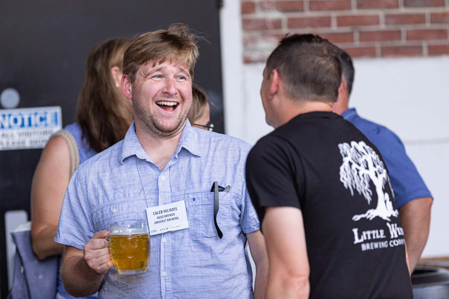 A man in a light blue shirt holds a glass mug of beer and laughs while talking with a man in a black Little Willow Brewing Company t-shirt, with a brick wall behind them.