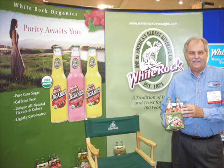A man in a blue shirt holds a pack of White Rock Organics soda beside banners for White Rock and White Rock Organics. A green director's chair and bottles in yellow and pink are nearby.