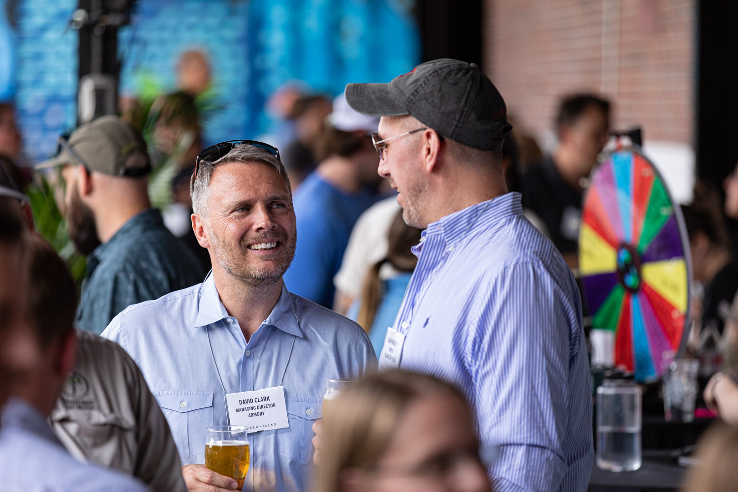 A smiling man in a light blue shirt with sunglasses on his head and a beer talks with a man in a gray cap and striped shirt, with a colorful prize wheel and the crowd behind them.