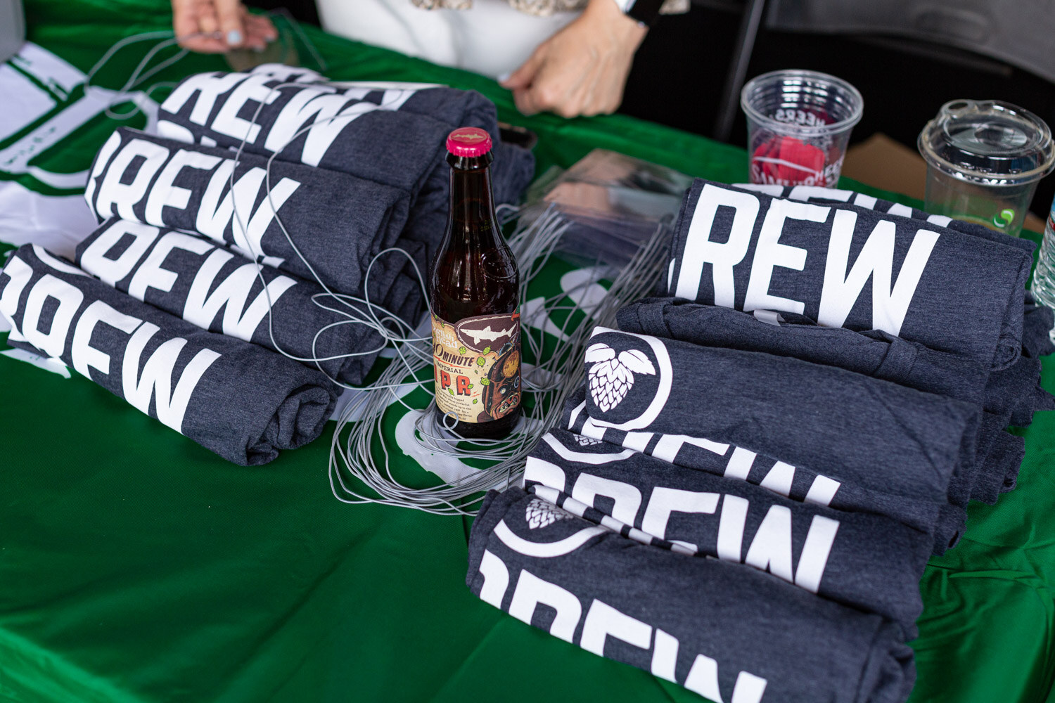 Close view of folded navy shirts printed BREW with a hop logo, loose lanyard cords, plastic cups and a bottle of Dogfish Head 90 Minute Imperial IPA on a green tablecloth.