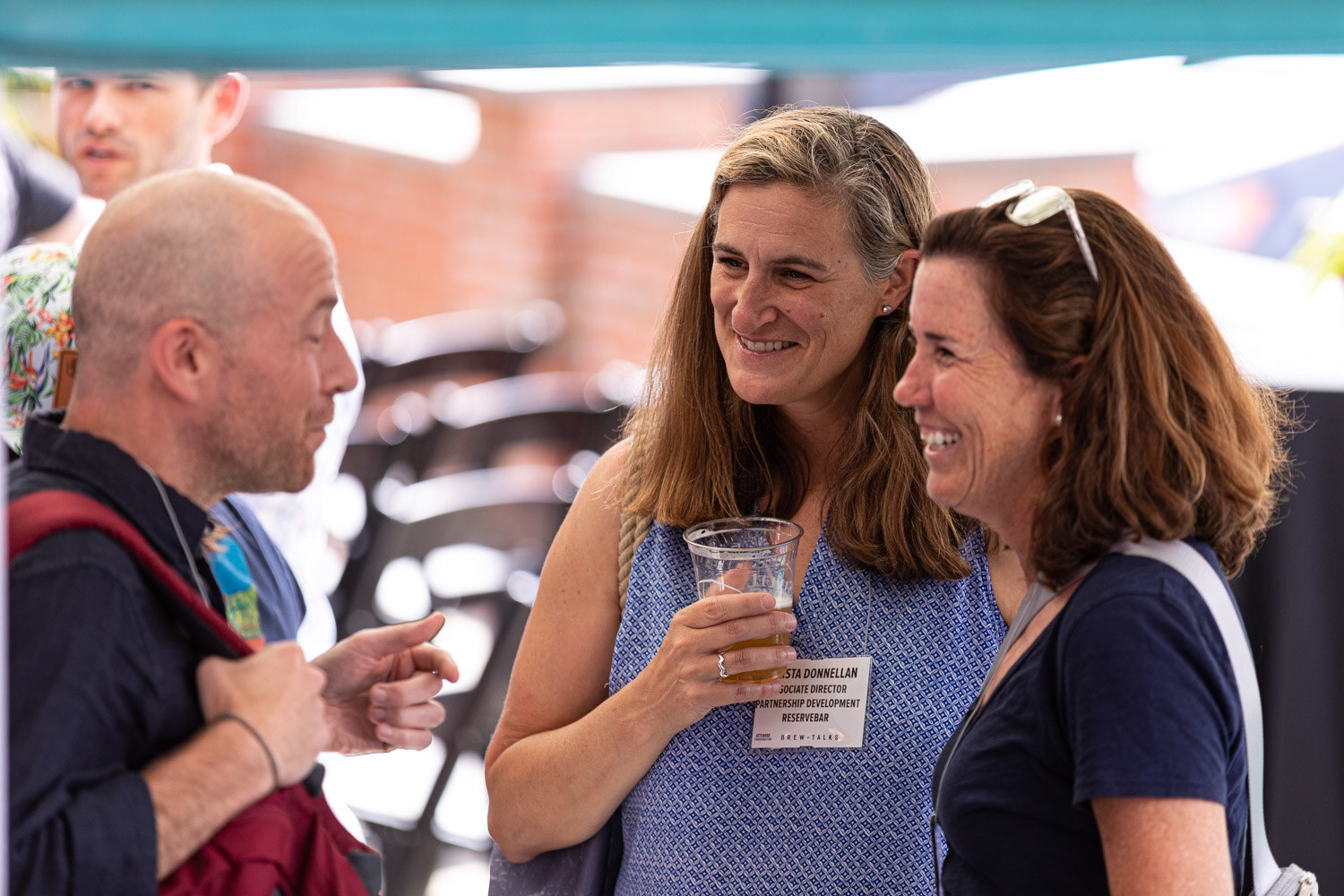 A smiling woman in a blue patterned top holding a cup of beer and a woman in a navy top with sunglasses on her head laugh while a bald man in a dark shirt and red backpack talks to them under a teal tent.