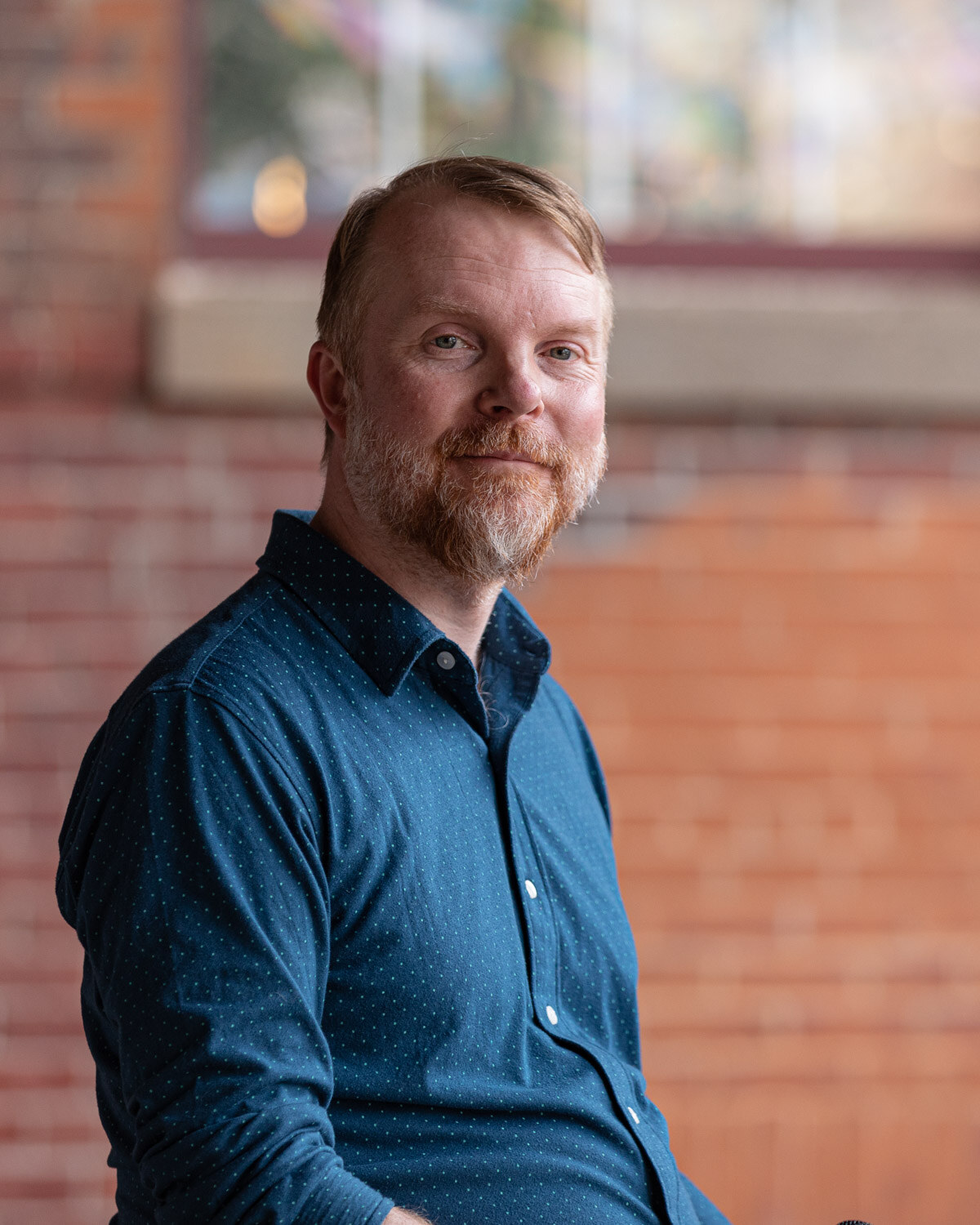 Portrait of a bearded man with thinning hair in a dark blue dotted button-up shirt seated in front of a red brick wall, looking at the camera with a slight smile.