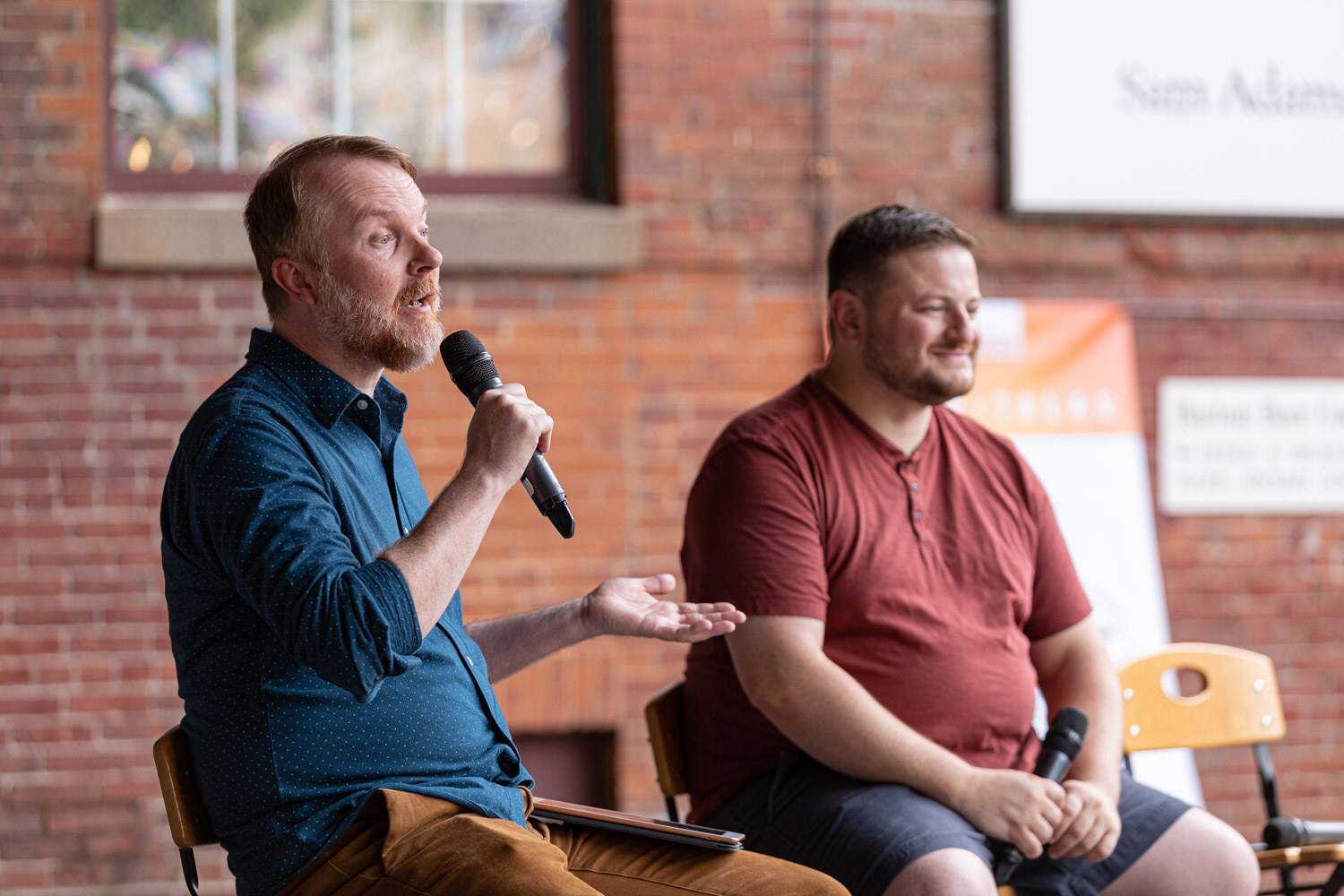 A bearded man in a dark blue dotted shirt speaks into a microphone with an open hand while a man in a maroon henley shirt and shorts smiles beside him, in front of a brick wall with a Sam Adams sign and an orange banner.