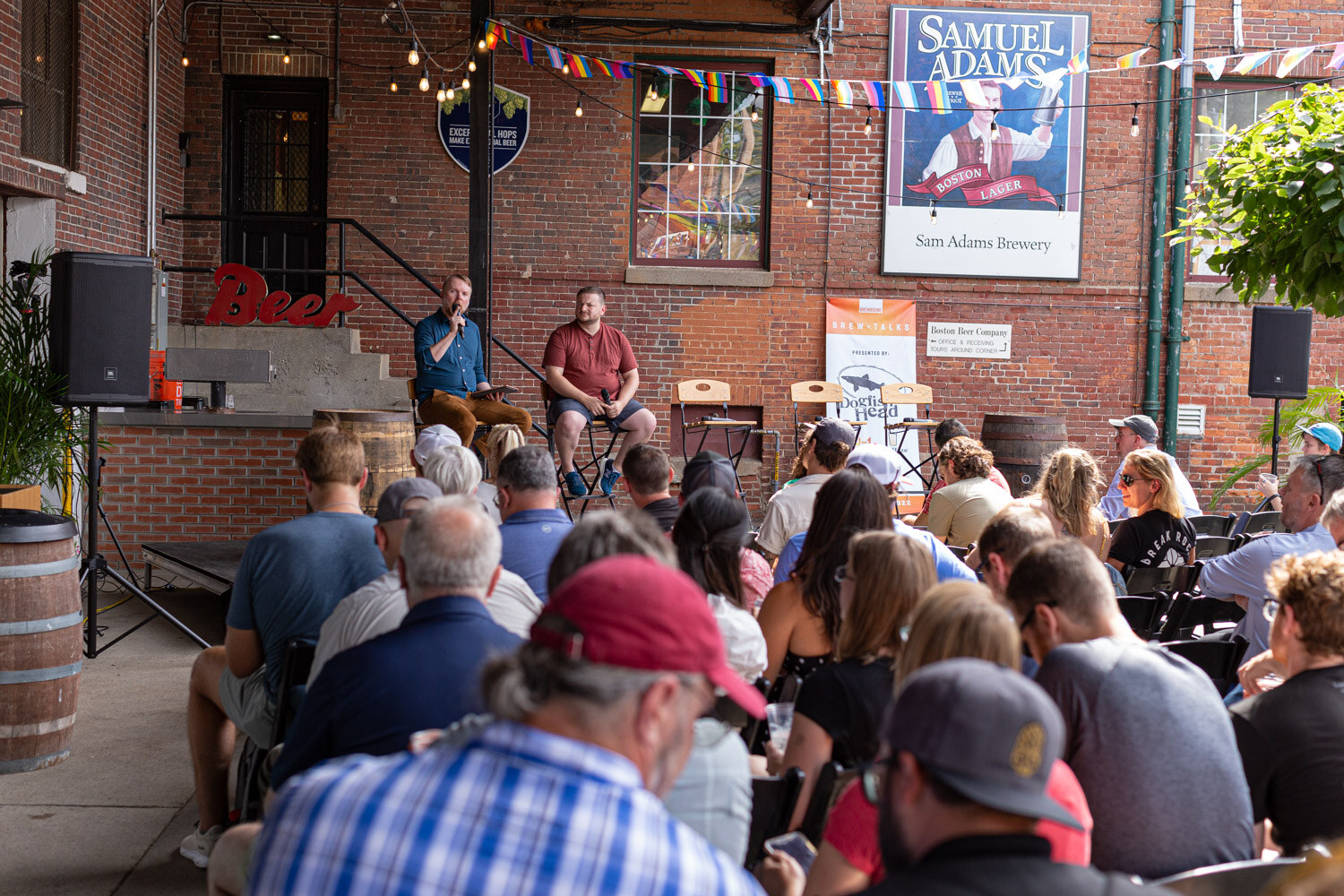 View over the heads of seated guests to two men talking on a small stage in front of a brick wall with a Samuel Adams Boston Lager sign, rainbow flags, string lights and a Brew Talks banner, with a barrel at the left.