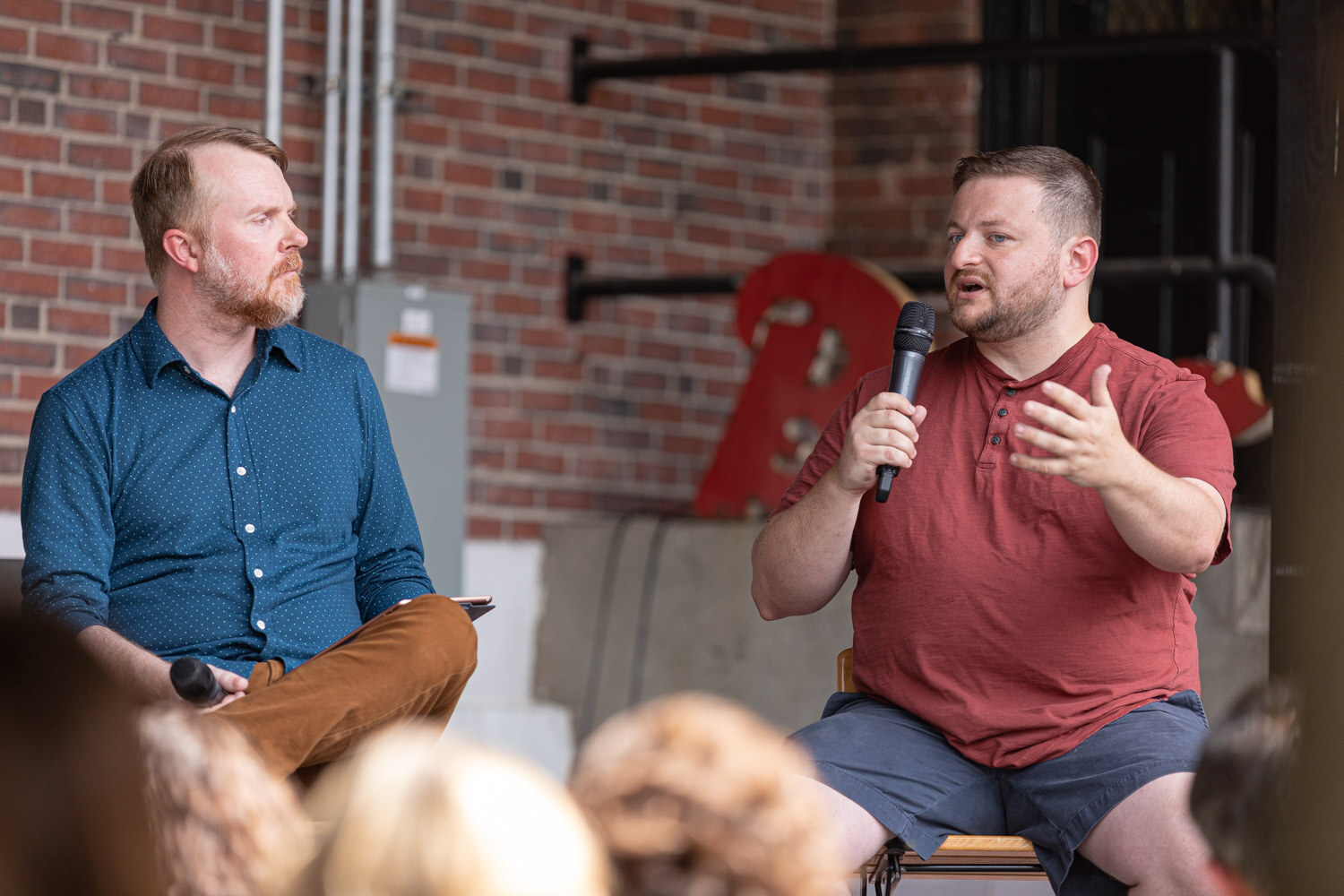 A man in a maroon henley shirt speaks into a microphone with an open hand while a bearded man in a blue dotted shirt looks on, in front of a brick wall with a red Beer sign, with audience heads in the foreground.
