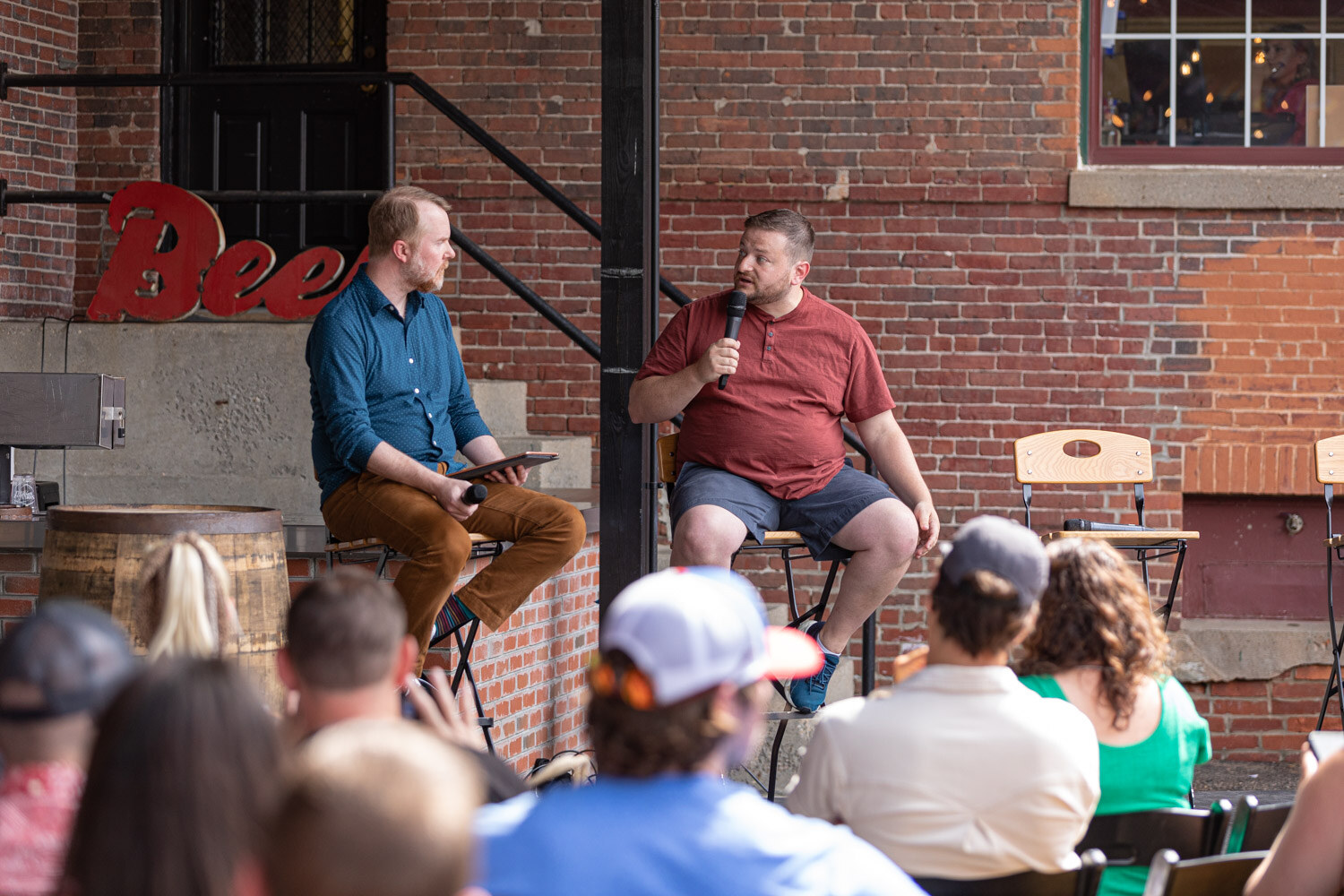 A bearded man in a blue shirt and tan pants holding a tablet and a man in a maroon shirt and shorts with a microphone face each other on stools in front of a brick wall with a red Beer sign, with the audience in the foreground.