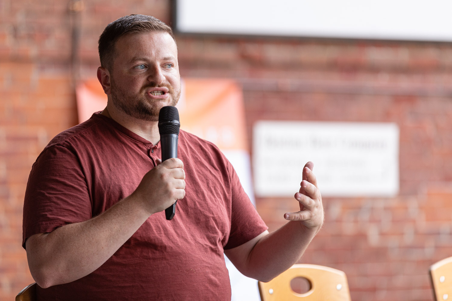 A man in a maroon henley shirt speaks into a microphone with one hand raised in front of a red brick wall with a blurred orange banner and white sign.