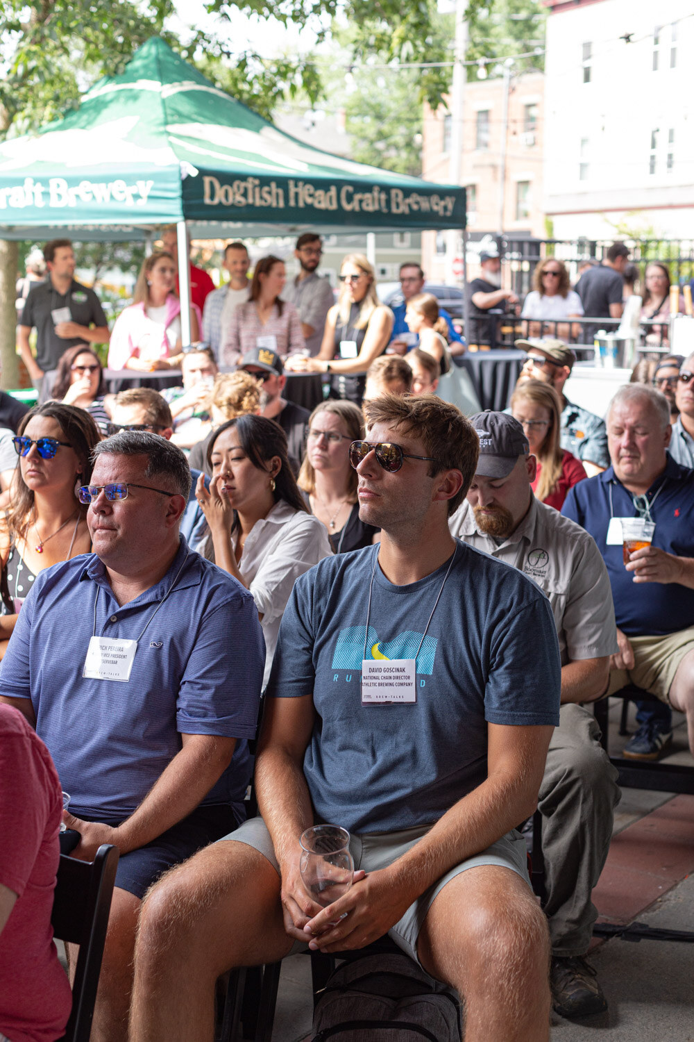 Close portrait-format view of seated attendees wearing name badges, including a man in sunglasses and a gray t-shirt and a man in a blue polo, with a green Dogfish Head Craft Brewery tent and standing guests behind them.