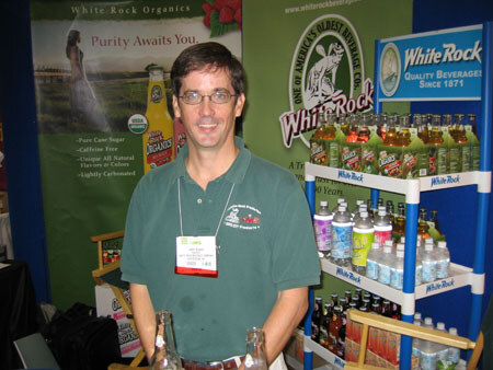 A man with glasses in a green polo shirt and a name badge stands in the White Rock Organics booth, with bottled drinks on blue shelves and a White Rock Quality Beverages sign beside him.