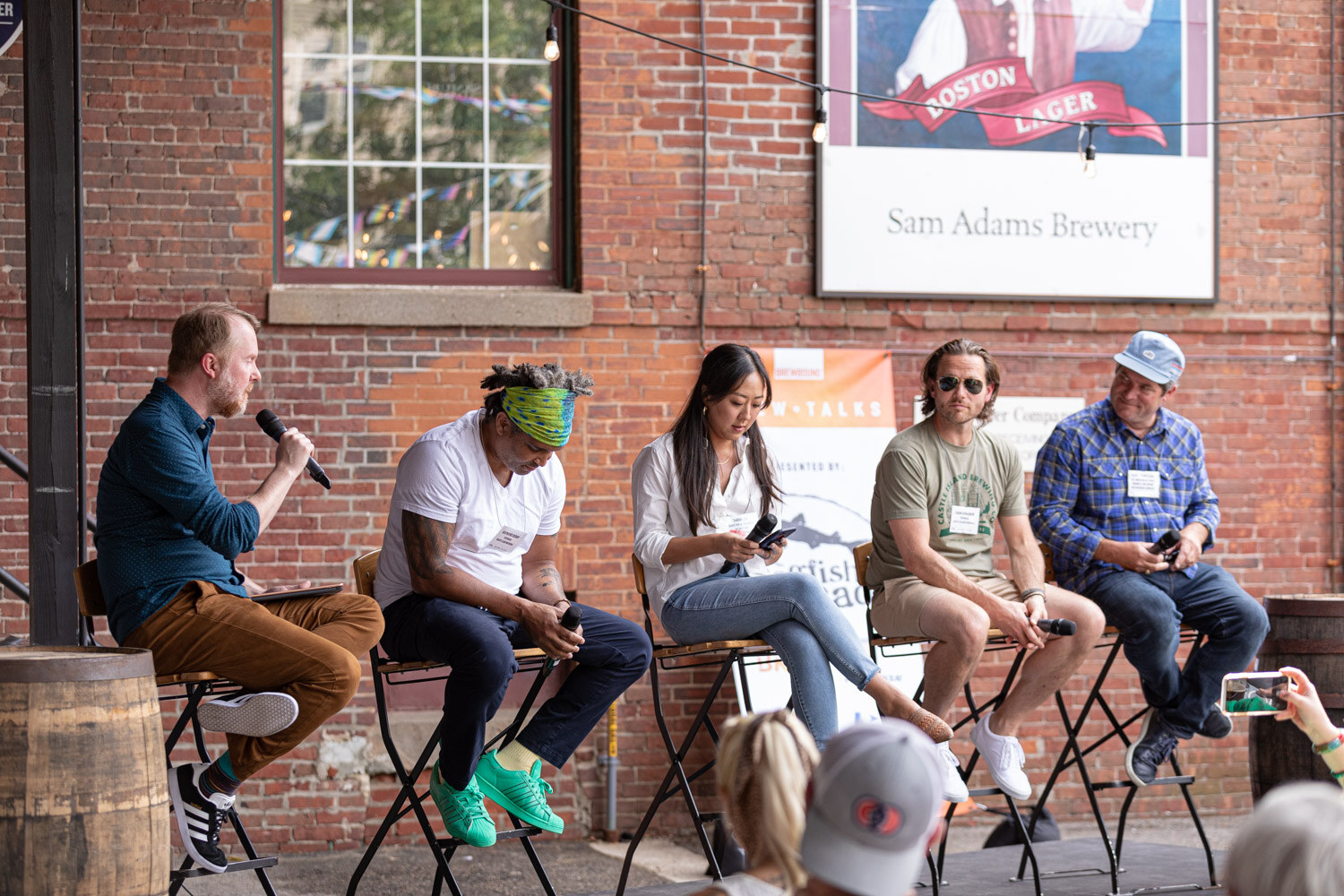 A bearded moderator in a blue dotted shirt speaks into a microphone beside four panelists on folding bar stools, a man in a green headband and white t-shirt, a woman in a white blouse with a phone, a man in sunglasses and an olive t-shirt, and a man in a blue plaid shirt and cap, in front of a brick wall and a Sam Adams Brewery sign.