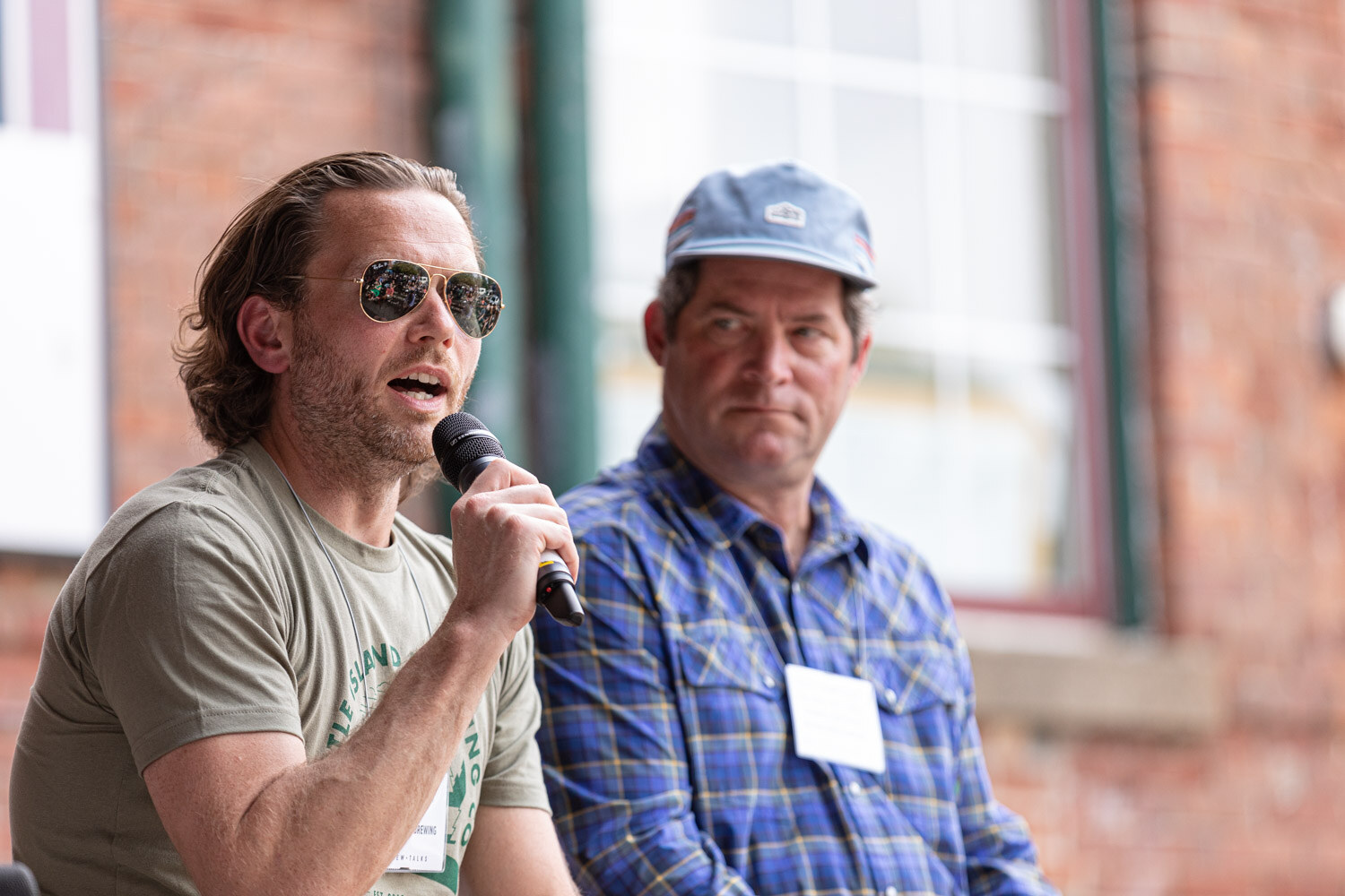 A man in aviator sunglasses and an olive graphic t-shirt speaks into a microphone while a man in a blue plaid shirt and light blue cap sits beside him, with a brick wall and a window at the back.