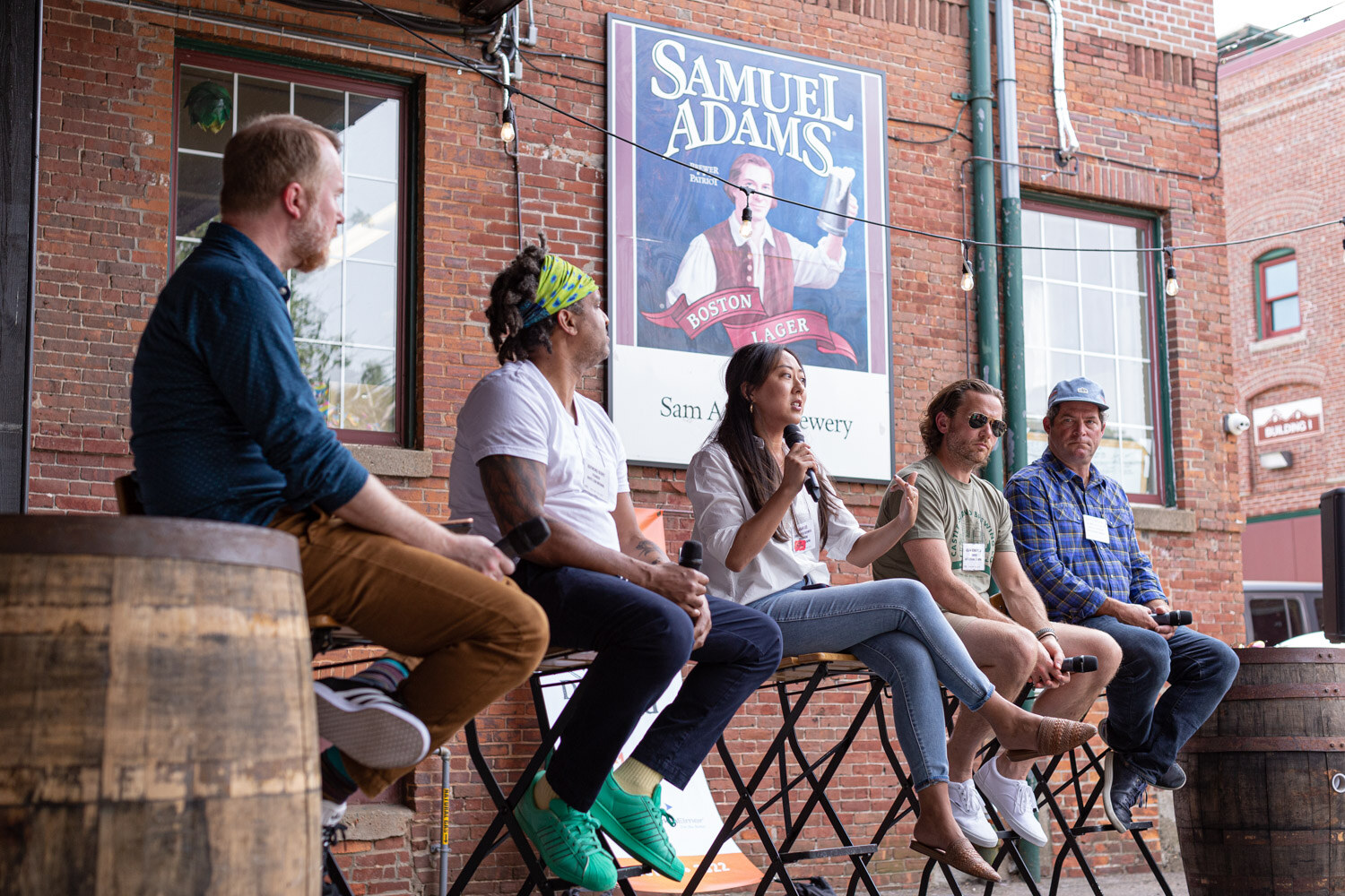 Low-angle side view of five people seated on folding bar stools in front of a brick wall under a Samuel Adams Boston Lager sign, with a whiskey barrel in the foreground at each side.