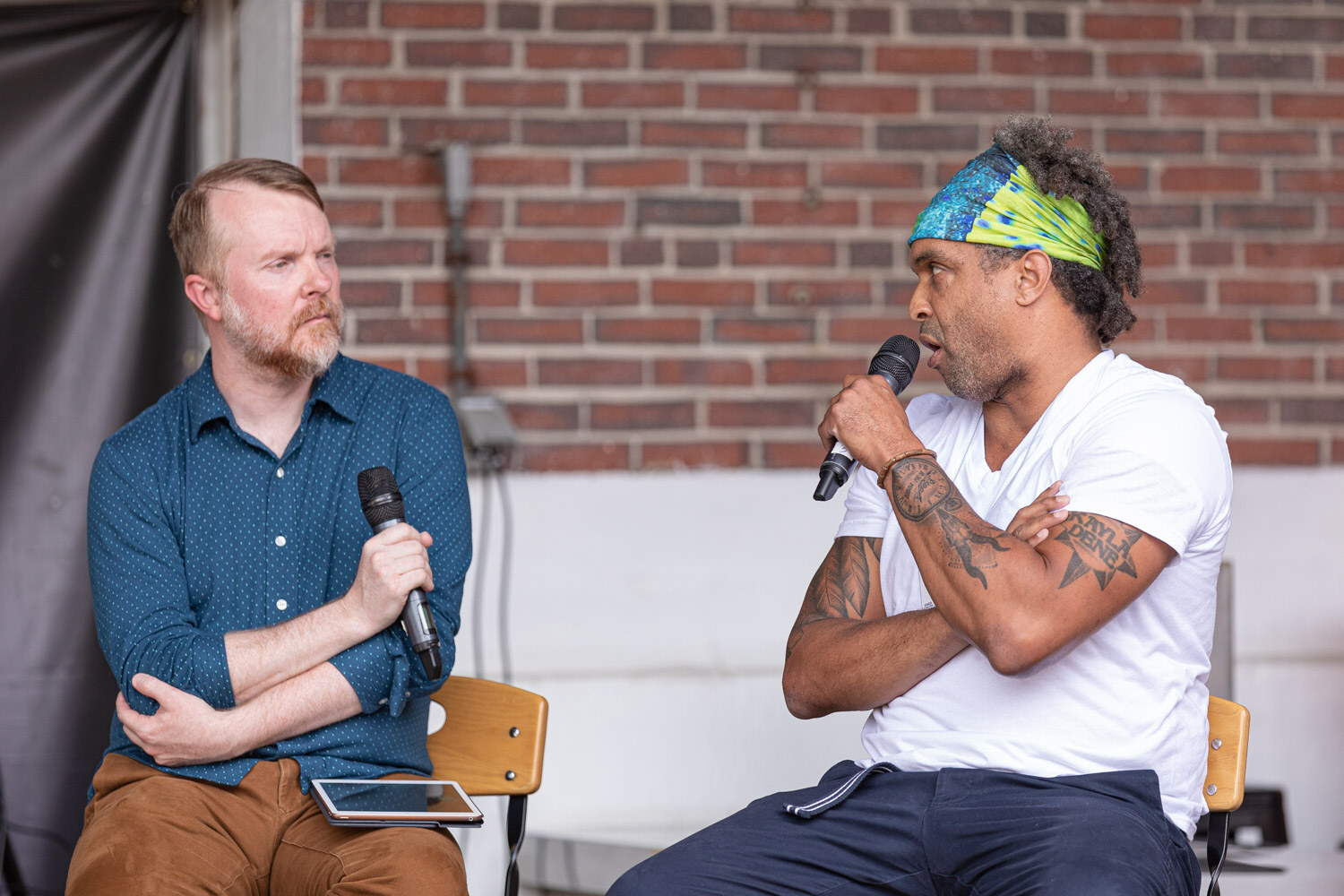 A bearded man in a blue dotted shirt holds a microphone and listens to a man in a green and blue headband and white t-shirt, who speaks into a microphone with his arms crossed, in front of a brick wall.