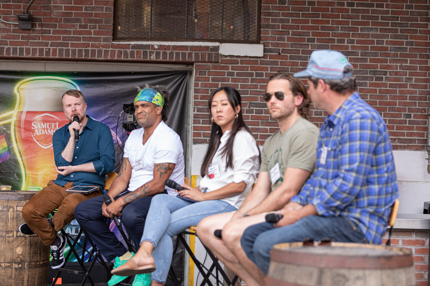 A bearded moderator in a blue shirt speaks into a microphone beside a man in a green headband, a woman in a white blouse, a man in sunglasses and a man in a plaid shirt and cap, seated in front of a black banner printed with a neon Samuel Adams glass.