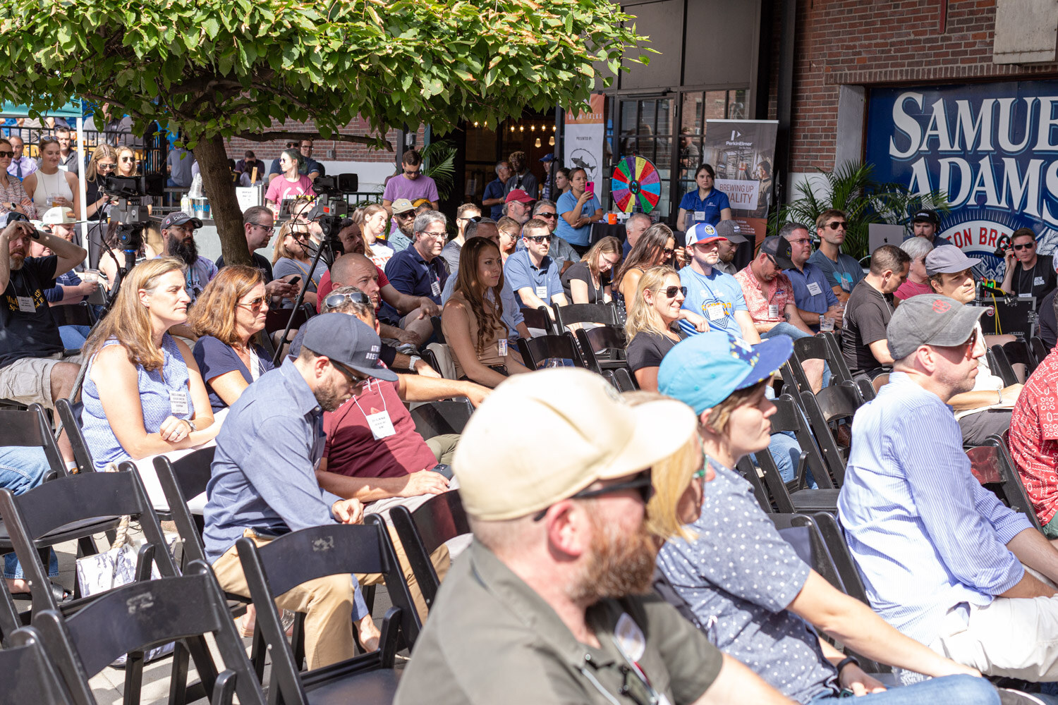 A packed audience of seated attendees with name badges in black folding chairs under a leafy tree, with a prize wheel and a Samuel Adams banner in the background.