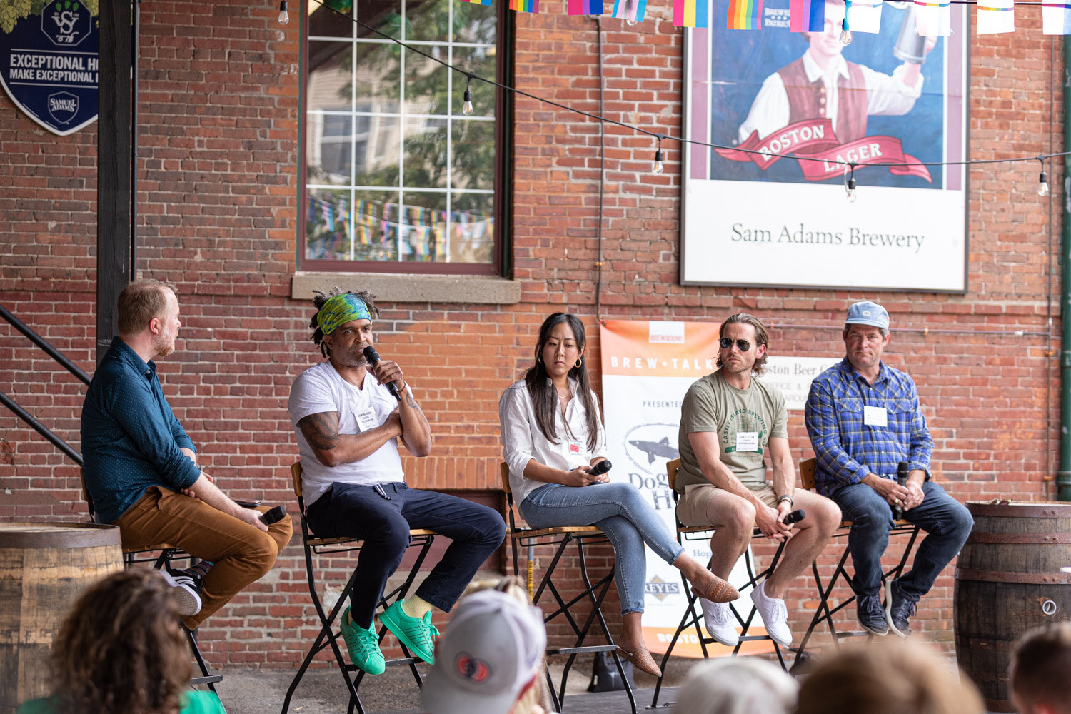 Five people on folding bar stools in front of a red brick wall with a Samuel Adams Boston Lager sign, string lights, rainbow pennant flags and a Brew Talks banner, with a whiskey barrel at each end and audience heads at the bottom edge.