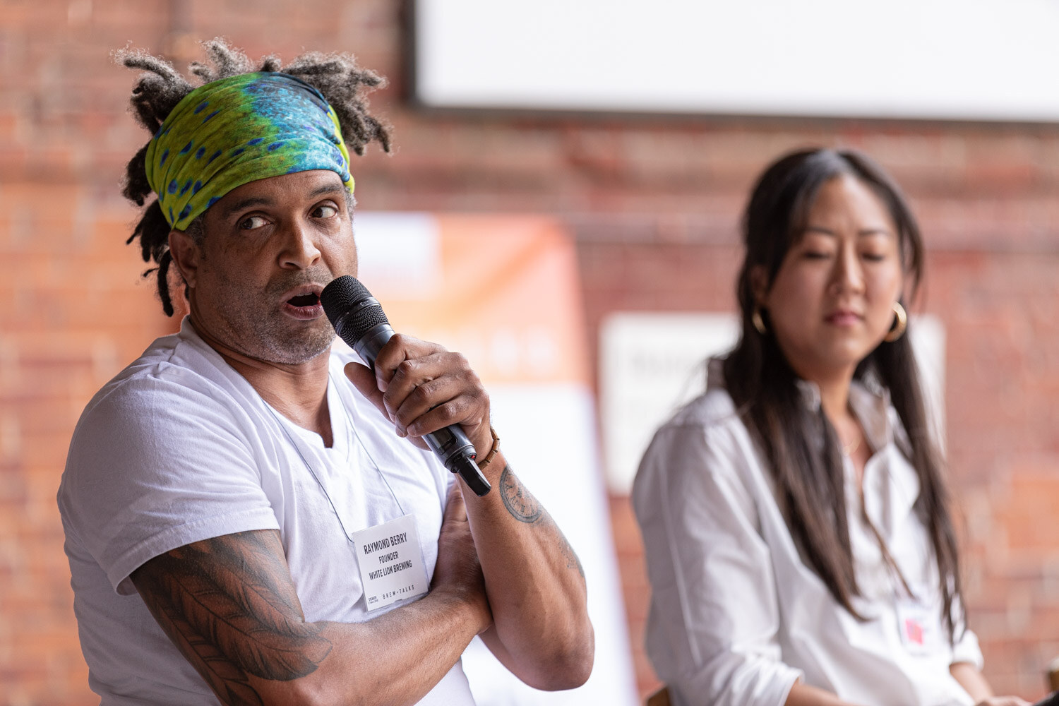 A man in a green and blue headband and white t-shirt speaks into a microphone while a woman in a white blouse sits out of focus behind him, in front of an orange banner and a brick wall.