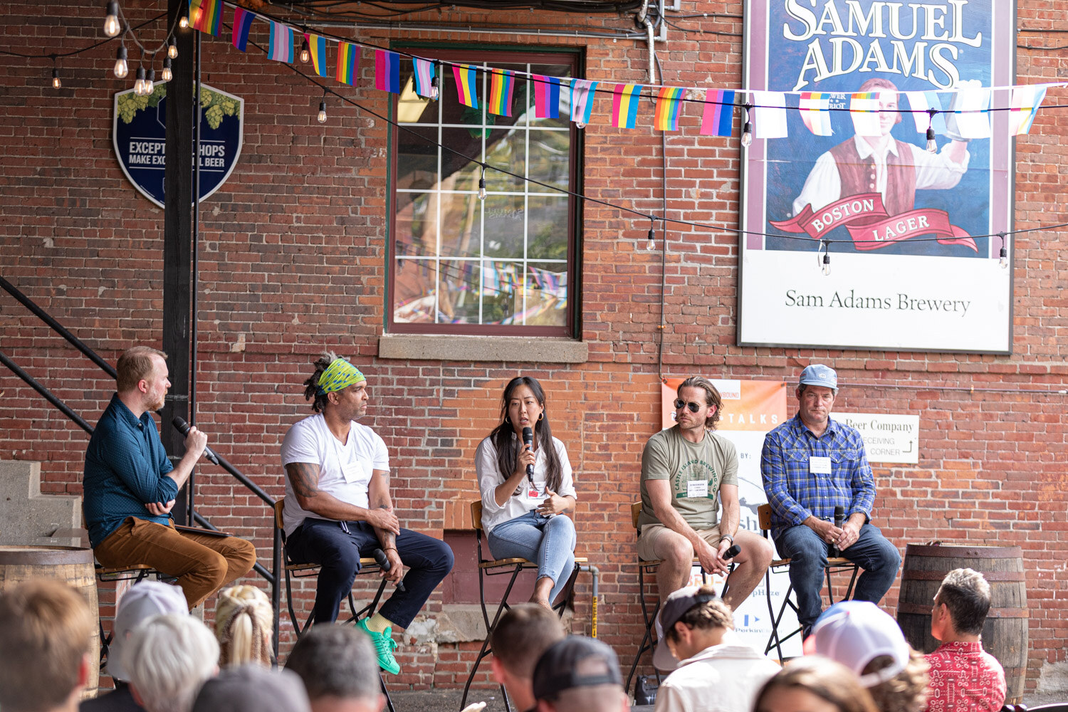 A woman in a white blouse speaks into a microphone while the moderator, a man in a green headband, a man in sunglasses and a man in a plaid shirt sit on stools in front of a Sam Adams Boston Lager sign, with the audience in the foreground.