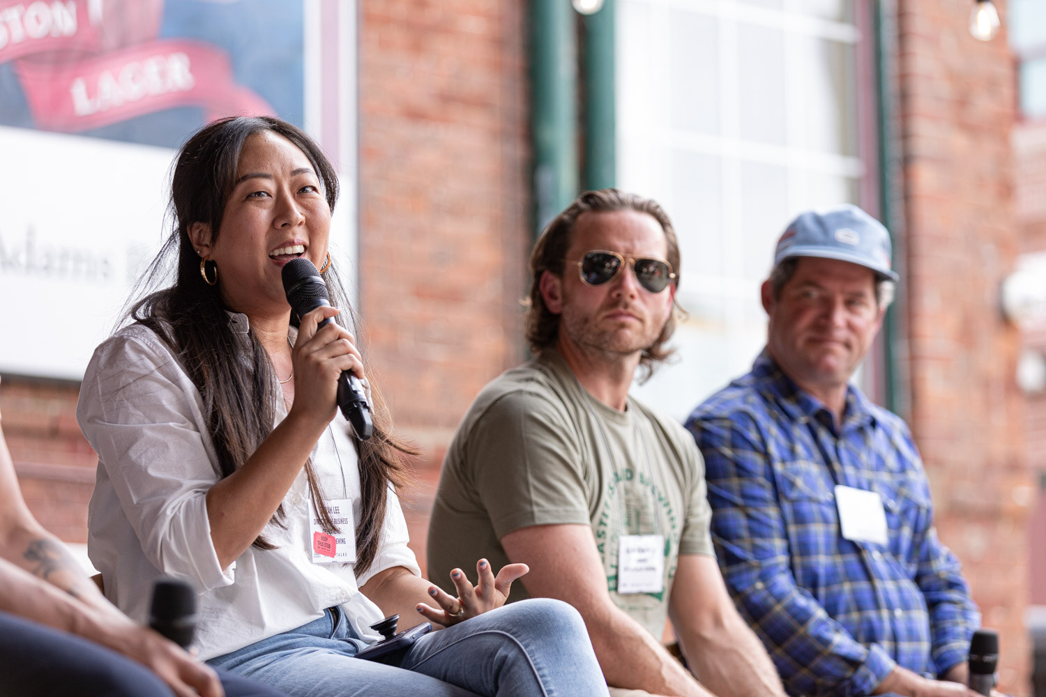 A woman in a white blouse and gold hoop earrings speaks into a microphone while a man in sunglasses and an olive t-shirt and a man in a blue cap and plaid shirt sit beside her.