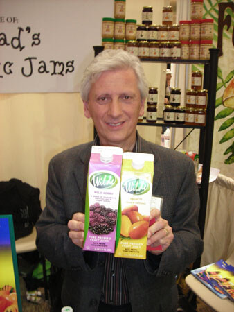 A gray-haired man in a dark jacket holds up two cartons of Wild Berry and Wild Mango pure pressed fruit juice. Jars of jam sit on shelves behind him.