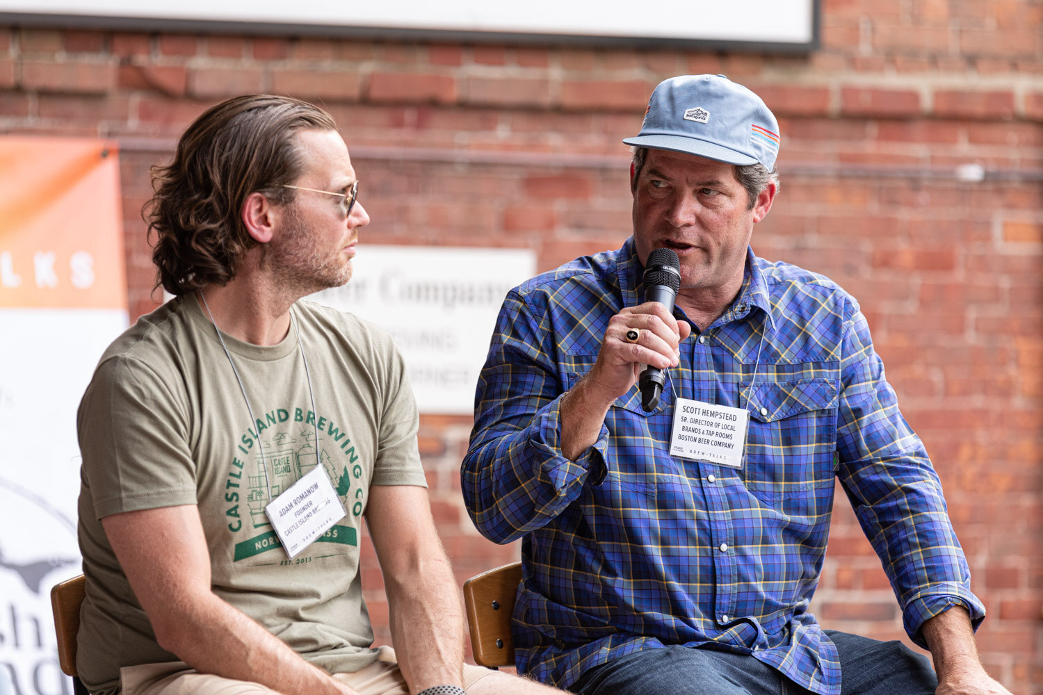 A man in a light blue cap and blue plaid shirt speaks into a microphone while a man in glasses and an olive Castle Island Brewing Co. t-shirt looks to the side.