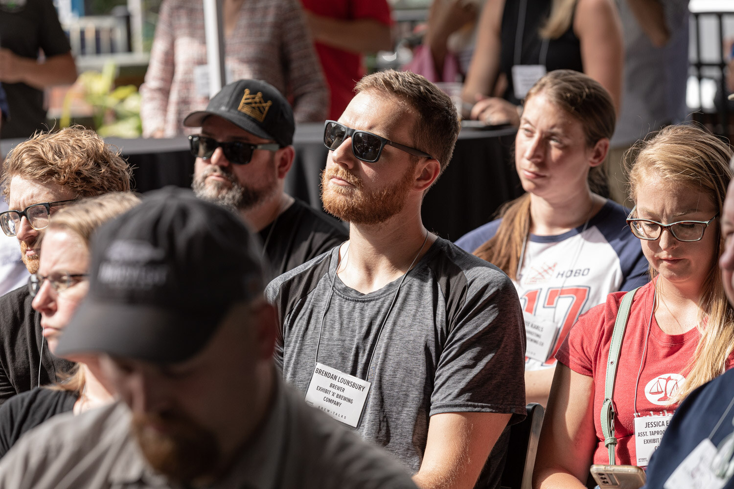 Close view of seated attendees with name badges, including a bearded man in sunglasses and a gray t-shirt, a man in a black cap, and a woman in a red t-shirt and glasses, looking toward the stage.