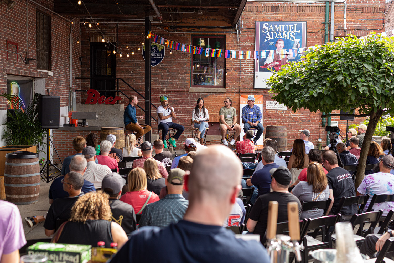 Wide view over seated guests to five panelists on stools in front of a brick wall under a Samuel Adams sign with rainbow flags, with a leafy tree at the right and a case of Dogfish Head beer in the foreground.