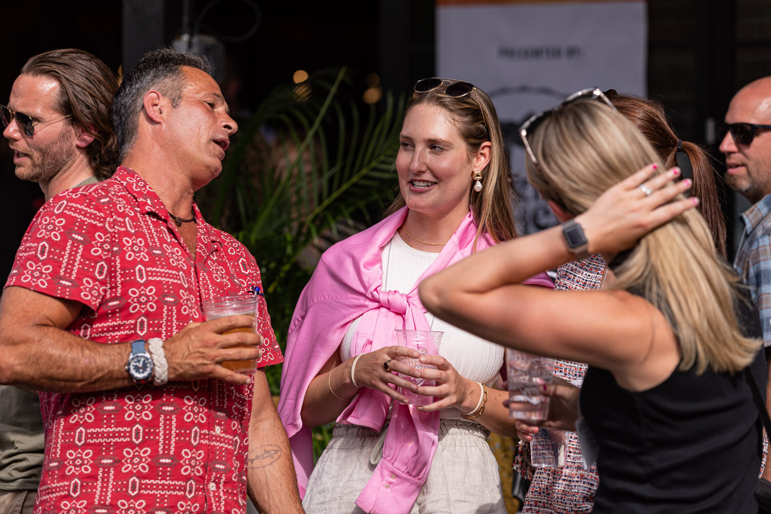 A man in a red patterned shirt with a drink talks near a smiling woman in a pink wrap and white top while a woman in black with sunglasses on her head runs a hand through her hair, with palms behind them.