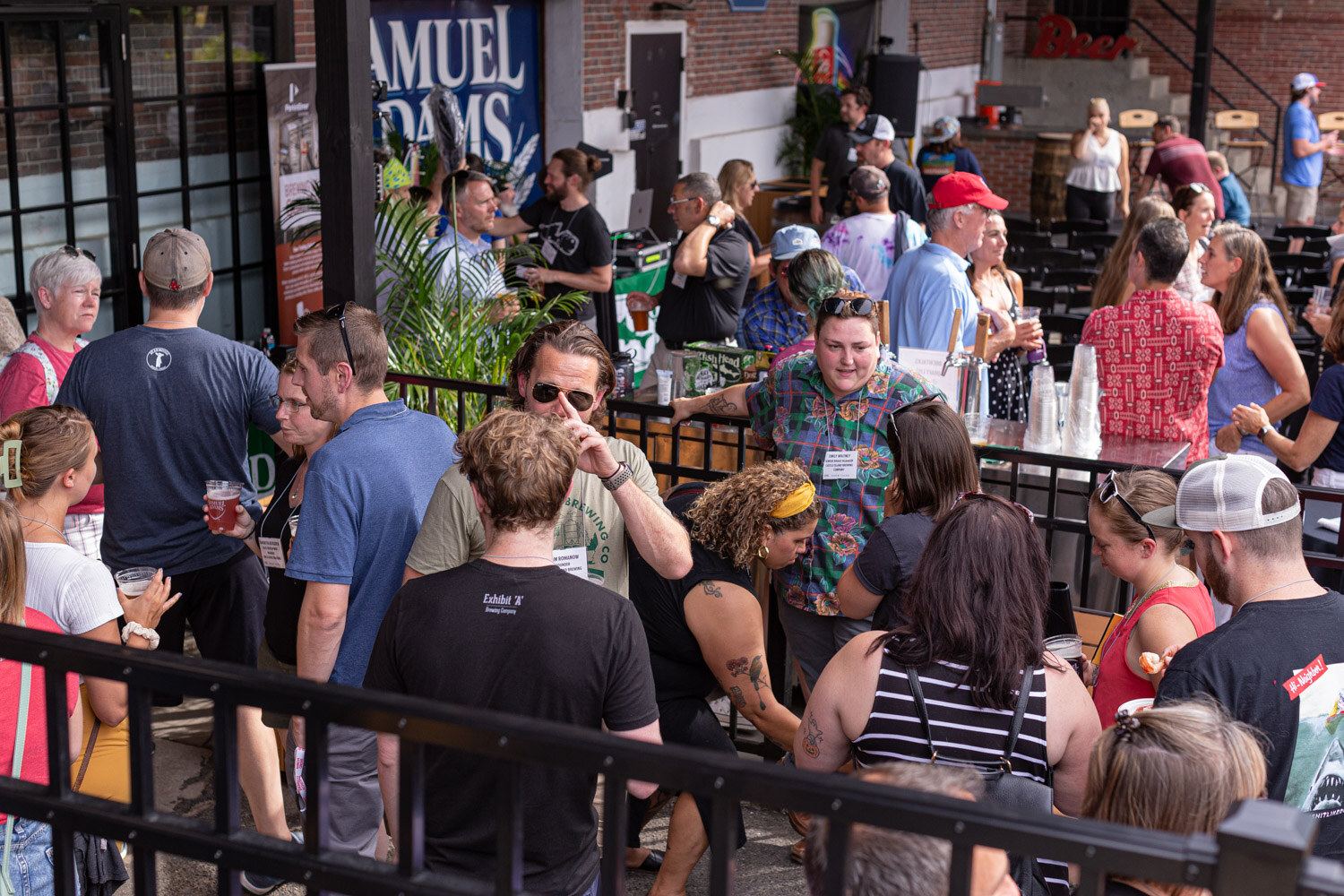 Elevated view of guests with beers crowded around a bar with taps behind a black iron railing, with palm plants, a Samuel Adams banner and a brick wall in the background.